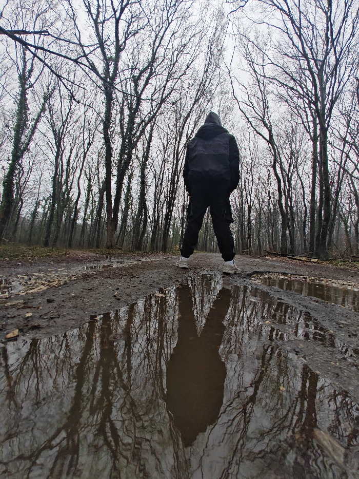 A single individual stands centrally, facing away from the camera, on a muddy dirt path within the Shumen Plateau Nature Park, Shumen, Bulgaria. The subject wears dark outerwear, including a hooded jacket and trousers, paired with light-colored athletic footwear. Their feet are positioned on the damp path, which features multiple large puddles in the foreground and midground. These puddles provide a clear reflection of the individual's silhouette and the surrounding leafless trees. The environment is characterized by a dense forest of tall, deciduous trees with bare branches, some exhibiting ivy growth on their trunks, extending towards an overcast, uniformly gray sky. The ground is consistently muddy and uneven, indicating wet conditions. The scene suggests a late autumn or winter day.