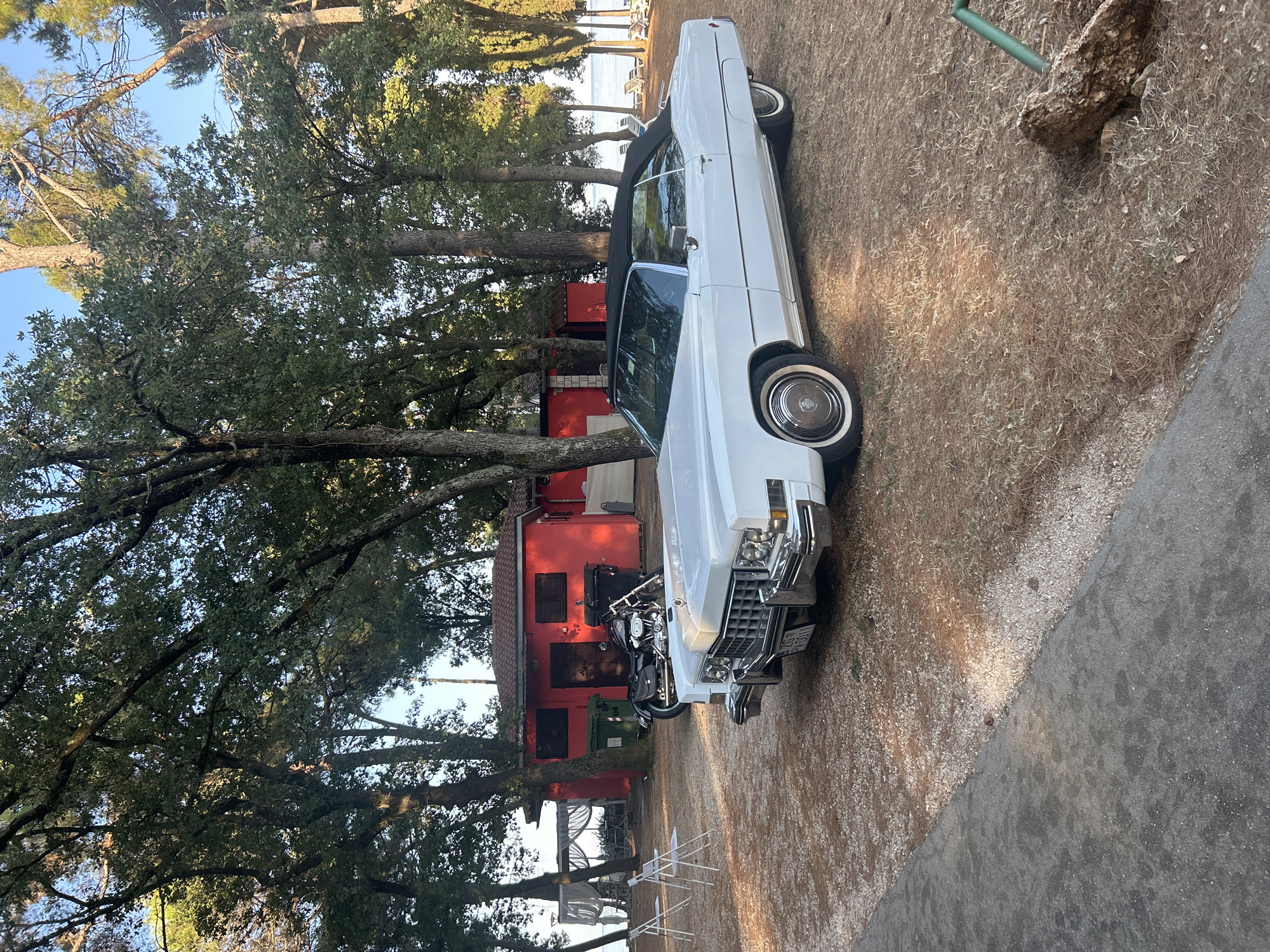 This image depicts a white convertible Cadillac parked on a gravel area in front of a red building. The Cadillac has its top down and classic whitewall tires. To the left of the Cadillac, a black motorcycle is parked near the red building. The building has two dark windows or shutters on its facade and appears to be a small structure, possibly a beach house or bar, given the presence of lounge chairs in the background near the water. Trees with dense green foliage surround the scene, casting shadows on the ground, which is covered with gravel and dry pine needles. The sky is clear and blue. The location is identified as Monterol, Croatia. There are no people visible in the image.