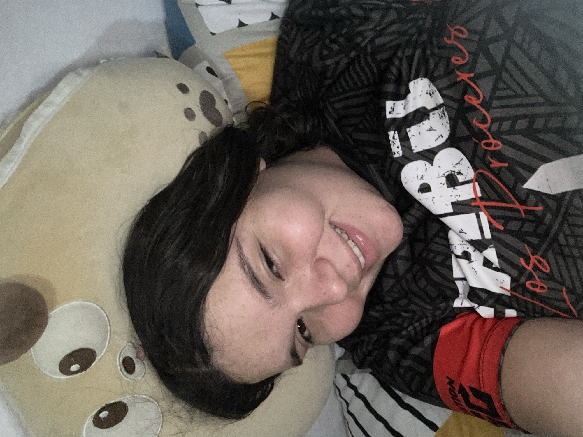 A young woman lies on her side on a bed, smiling slightly at the camera. Her head rests on a plush, light-brown animal pillow with darker brown spots. She is wearing a dark grey t-shirt with black geometric patterns and a white and red print of the word "Procesos". Red athletic shorts with dark lettering on the waistband peek out from beneath the t-shirt. The bed has a striped pillowcase and a patchwork quilt. The scene's location is unknown; there is no evidence suggesting San Juan de Los Morros, Venezuela.