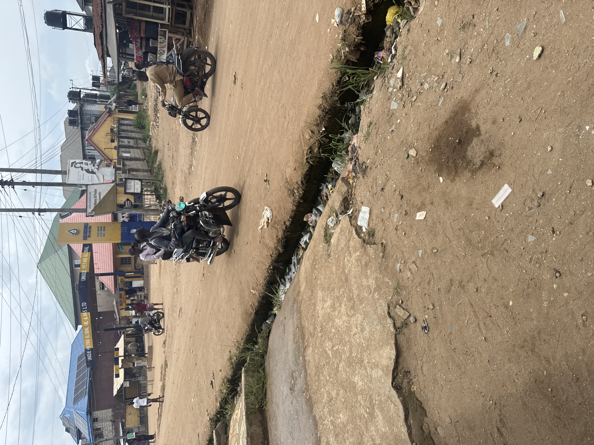 The image shows a street scene in Ihiagwa, Nigeria. The road is mostly dirt, with a narrow strip of cracked concrete on one side. A deep, garbage-filled gully runs along the edge of the road. Motorbikes are the primary mode of transportation, with several people riding them along the road. Buildings and businesses line the street, including a "UNIQUE PETANI GLOBAL OIL & GAS LTD" gas station with solar panels on its roof. There are also visible power lines and utility poles, plus water storage tanks on top of a building. The weather appears overcast.