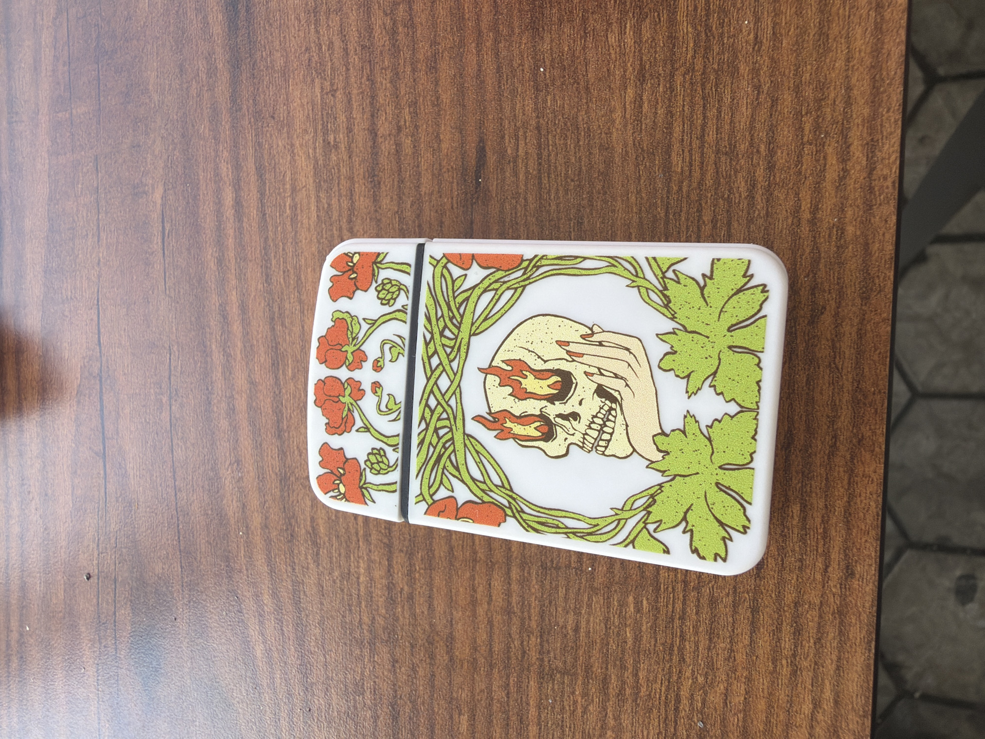 A white, rectangular object, possibly a lighter or small case, lies horizontally on a reddish-brown wooden surface. The object features an elaborate artistic design with a central motif of a skull. Flames emanate from the skull's eye sockets, and a hand with red nail polish partially covers one of its eyes. Surrounding the skull and hand are intricate green leafy vines and vibrant red-orange flowers, creating a decorative border. A thin black line divides the object's surface, indicating a potential hinge or seam. The wooden surface displays a visible grain pattern, and to the right, a section of a grey tiled floor with a hexagonal pattern is visible. The scene is brightly lit, suggesting an indoor setting during the daytime in Borovo, Bulgaria. No text is visible on the object or in the background. The design elements evoke a blend of gothic and natural aesthetics.