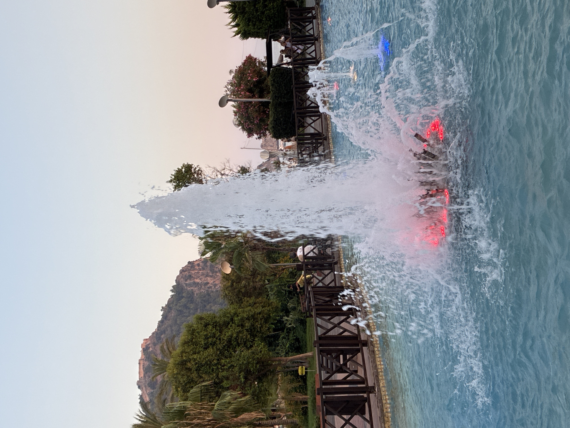 The image captures a fountain in the city of Alanya, Turkey, with a castle on a hill in the background. The fountain is active, with water spraying upwards and outwards, illuminated by red and blue lights at its base. In the midground, there is a wooden fence bordering a pathway, and beyond that, several people are visible sitting or walking. Palm trees and other greenery are scattered throughout the scene, and the sky is a clear, pale blue with a hint of pink near the horizon, suggesting it is either early morning or late evening. The water in the fountain is a vibrant blue.
