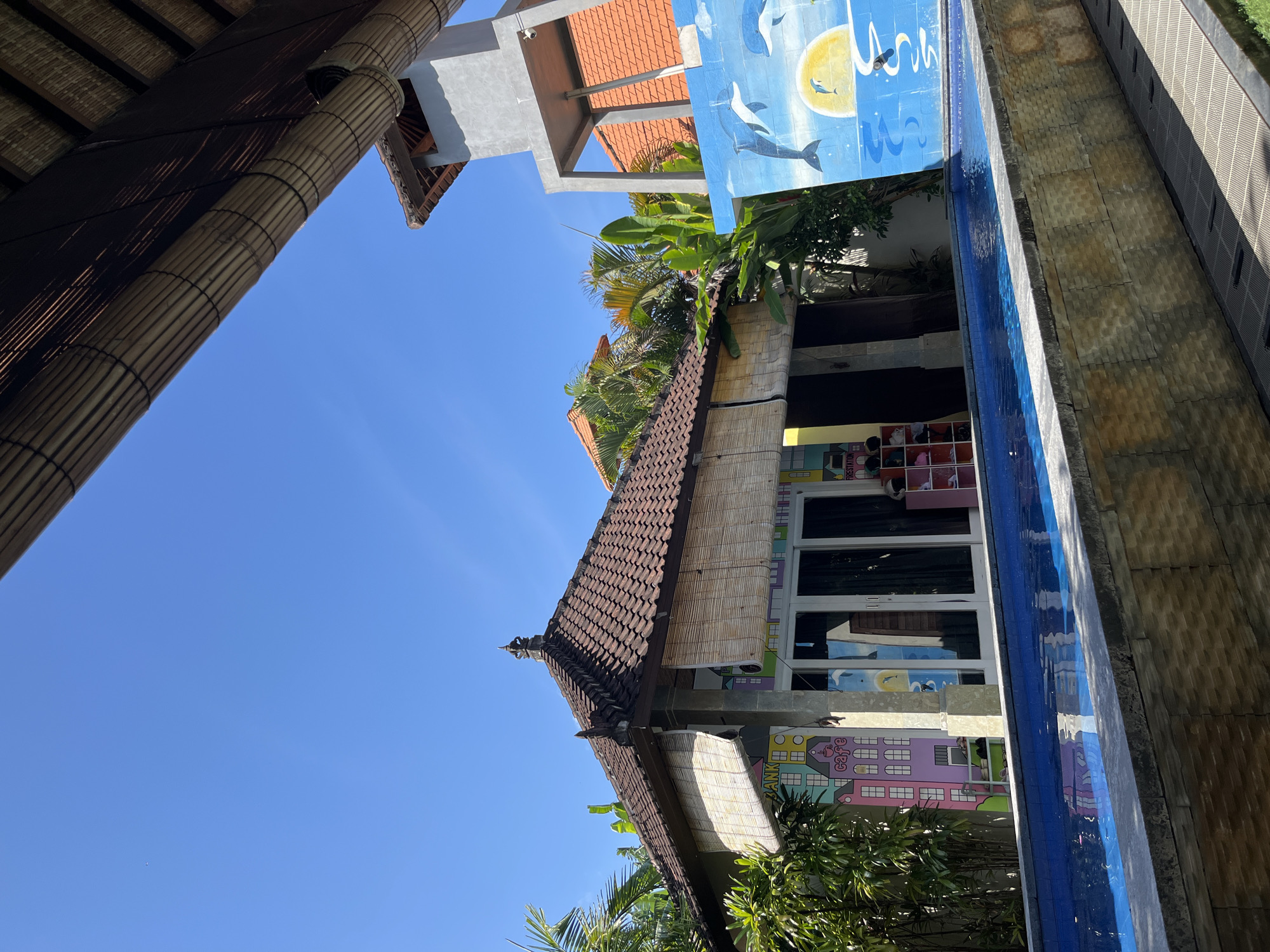 The image shows a building in Kuta, Indonesia, under a clear blue sky. The building has a brown tiled roof with a decorative finial. There are bamboo blinds partially covering the facade. The building's wall is decorated with colorful murals, including cityscapes and marine life. A swimming pool runs along the side of the building, reflecting the blue sky and murals. Parts of the building are seen through trees.