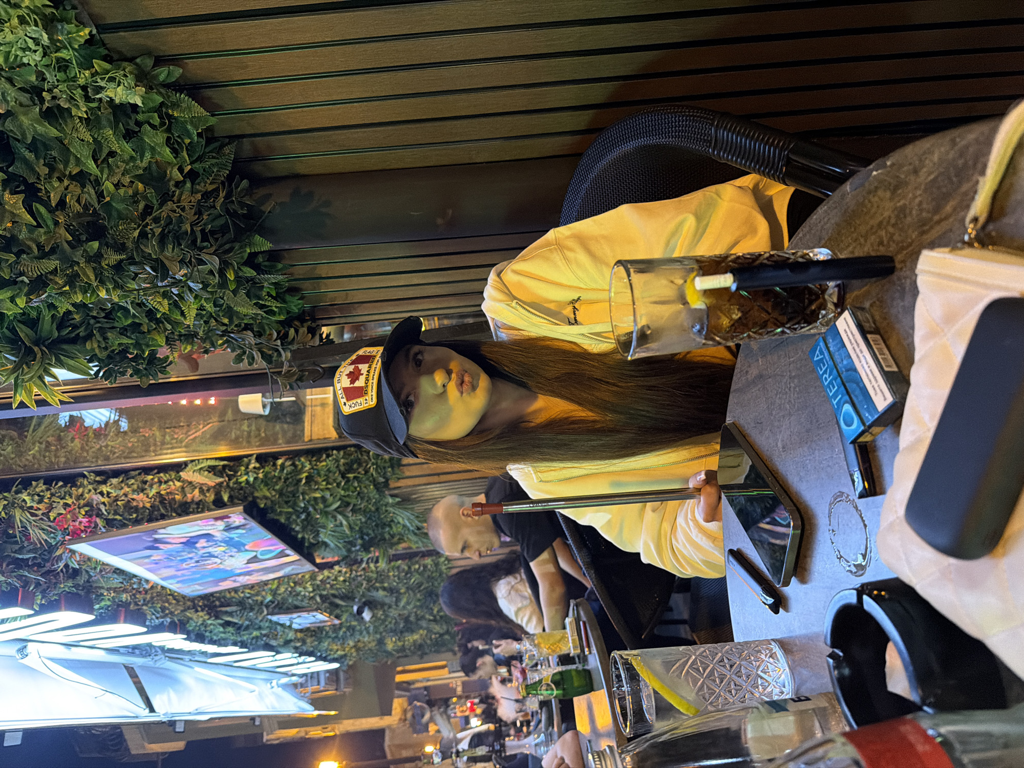 The image shows a woman sitting at an outdoor table in what appears to be a restaurant or bar in Unknown, Unknown. The woman is wearing a black cap with a Canadian flag patch, a light yellow hoodie, and has long brown hair. She is holding a hookah hose in her hand. On the table, there's a glass of liquid, a smartphone, a pack of cigarettes, a lighter, a purse, and various other bar-related items such as empty glasses and bottles. Other people are visible in the background, sitting at tables and engaging in conversations. The outdoor space is decorated with greenery and artificial lighting.