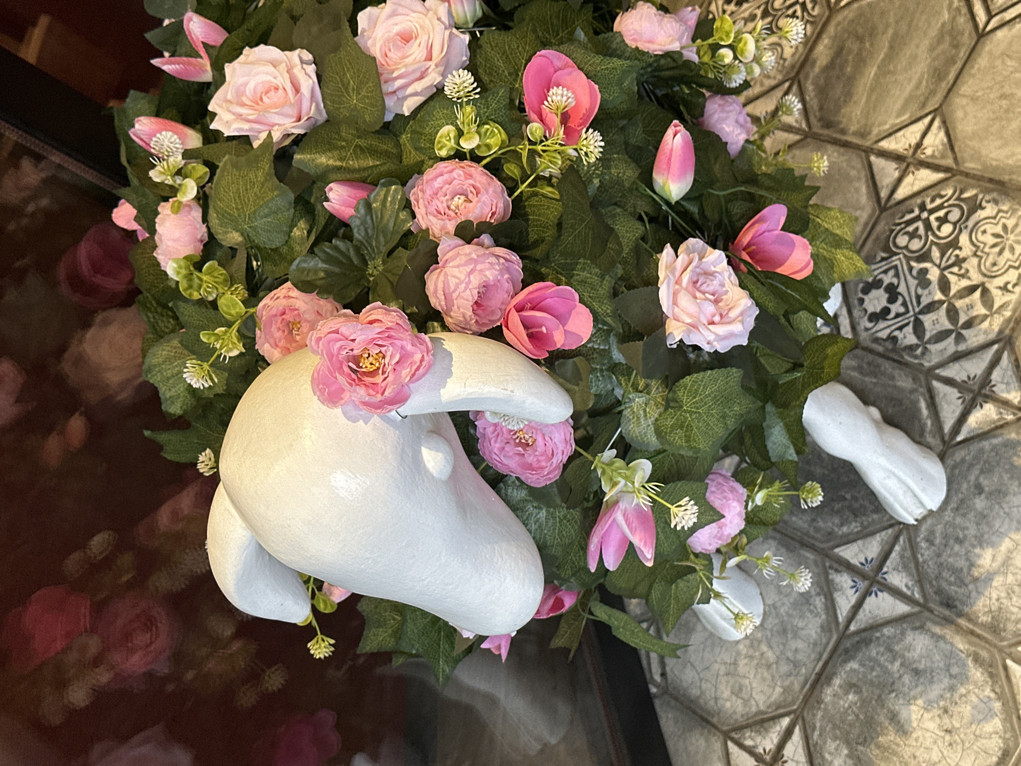 The image shows an overhead view of an ornamental arrangement placed either indoors or in a semi-indoor space. At the center is a stylized white sculpture, possibly made of plaster or a similar material. It is shaped like an animal head. Surrounding the white sculpture and filling the frame are artificial flowers, primarily pink roses and tulips, interspersed with green foliage and small white wildflowers. The arrangement sits partially on a surface that appears to be either a dark, reflective tabletop or a pane of glass reflecting a dark background. The other half is positioned on a tile floor with a geometric pattern in gray and white. The overall impression is that of an artistic display or a decorative centerpiece. There is no evidence to support the location of the city of Izhevsk, Russia.