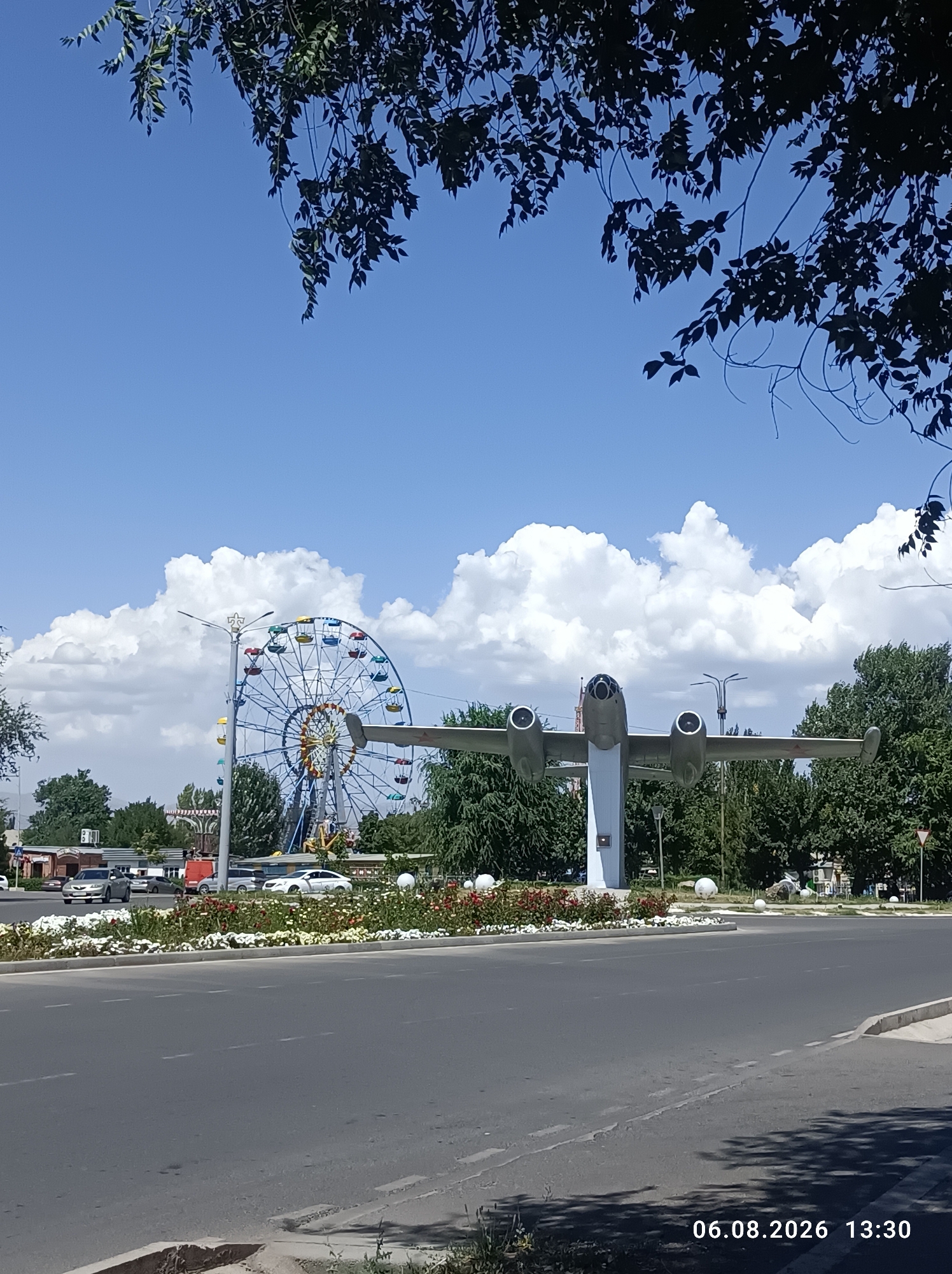 In Tokmok City, Kyrgyzstan, a monument of an airplane is displayed prominently in an outdoor setting. The airplane, a stationary, weathered monument, is mounted on a white pedestal and positioned in front of a large Ferris wheel and a lush green tree line. A road with white dashed lines runs through the foreground, with several cars parked or driving on it. The sky above is a clear blue, dotted with fluffy white clouds. The scene is illuminated by bright sunlight, casting shadows across the road and surrounding area. In the bottom right corner, a timestamp indicates the image was taken on August 6, 2026, at 1:30 PM.