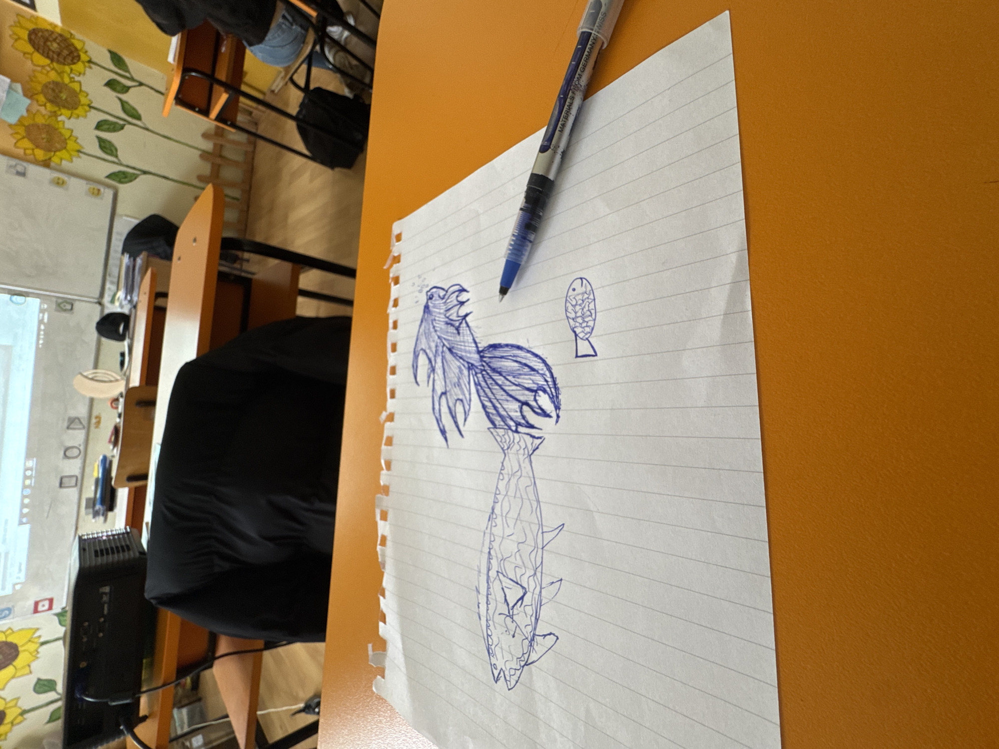 This media file depicts a classroom or study environment in Buhovo, Bulgaria. In the foreground, a lined piece of white paper rests on an orange-brown desk. On the paper, there are blue ink drawings: a detailed, stylized creature resembling a mythical fish or a bird-like entity with prominent fins and a wide-open mouth, and a smaller, simpler drawing of a fish. A blue ballpoint pen, partially labeled "MATERIALS FROM GERMANY SWISS," lies across the paper.
The background shows multiple wooden desks and chairs. A person's legs in jeans are visible under a desk in the upper left, and a black bag is stowed under another chair. The walls are light-colored and adorned with large, yellow sunflower decorations. A whiteboard is visible on the left wall, and a screen or projector displays a digital interface with various geometric icons (squares, circles, triangles). A black computer tower or electronic device is on the floor. The scene suggests an ongoing class or a break, with someone having engaged in drawing.