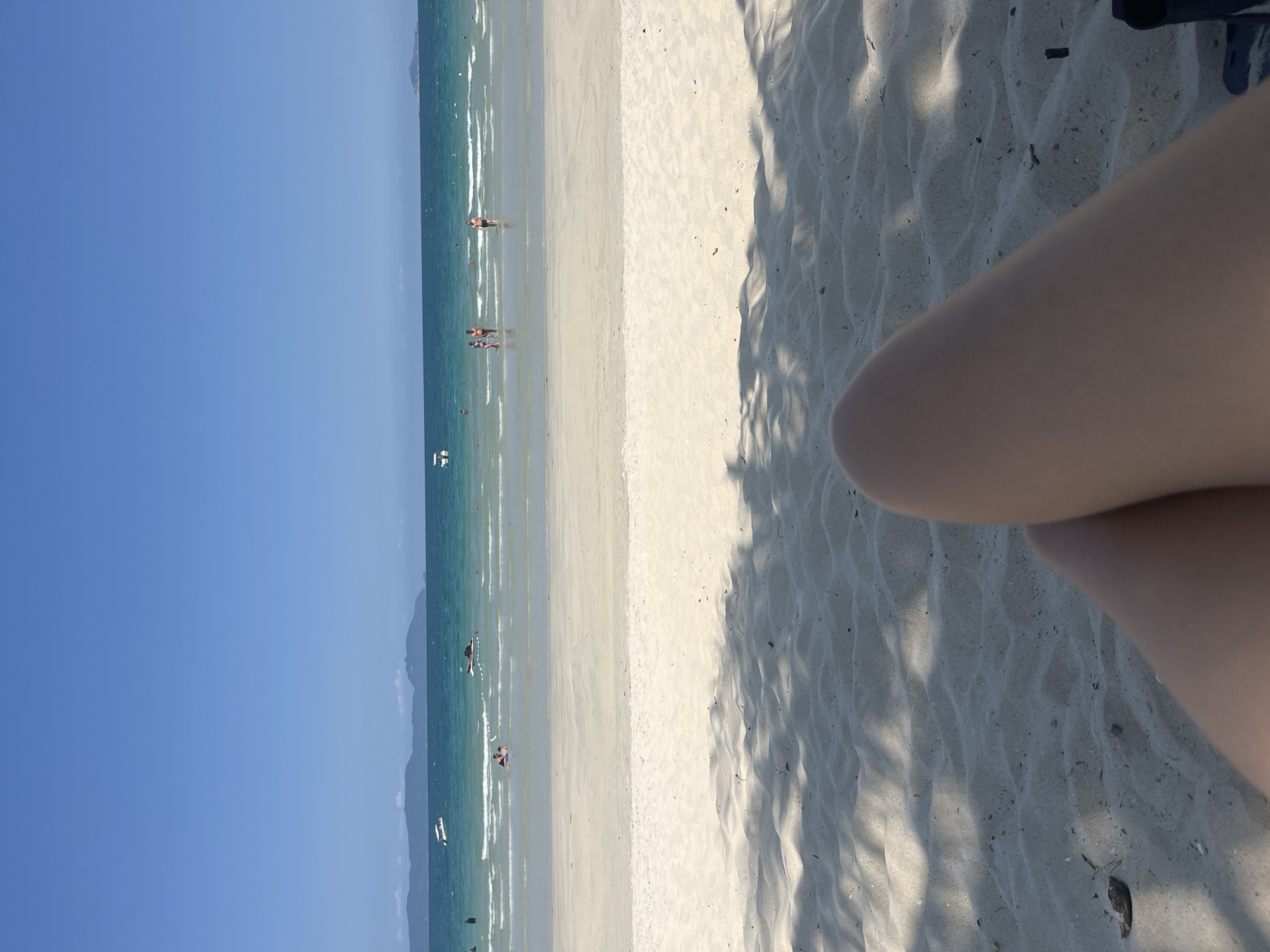 The image depicts a beach scene. In the distance, there is a clear blue sky meeting the ocean. Several people are visible in the water along the shoreline. The beach itself is sandy and divided into sections of direct sunlight and dappled shade, likely from trees. In the foreground, a person's leg is prominently visible, suggesting the image was taken from a personal perspective. The location is unknown, Unknown.