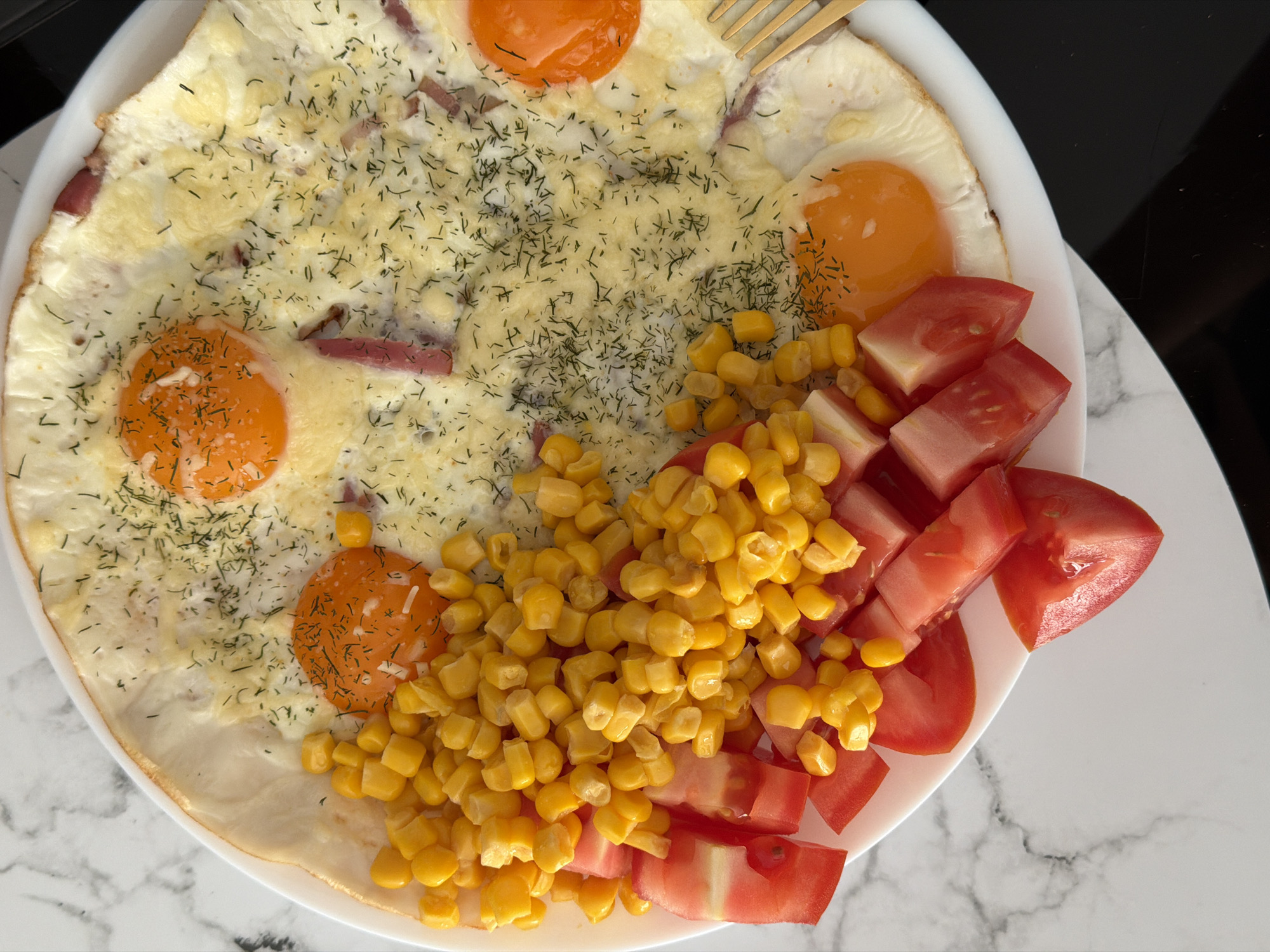 The image shows a close-up, overhead view of a breakfast dish on a white plate. The dish is arranged in sections. One section consists of diced red tomatoes. Adjacent to the tomatoes is a pile of cooked corn kernels. The largest section of the plate contains a fried egg dish with multiple sunny-side-up eggs, their bright orange yolks visible. The whites of the eggs are spread thinly, and there are pieces of what appear to be ham or sausage mixed in. The egg dish is sprinkled with chopped dill. A gold fork is partially visible on the right side of the plate. The plate rests on a surface with a white and gray marble pattern. The overall setting appears to be an indoor environment, likely a kitchen or dining area, but no specific location in Finestrat, Spain, can be determined from the image.