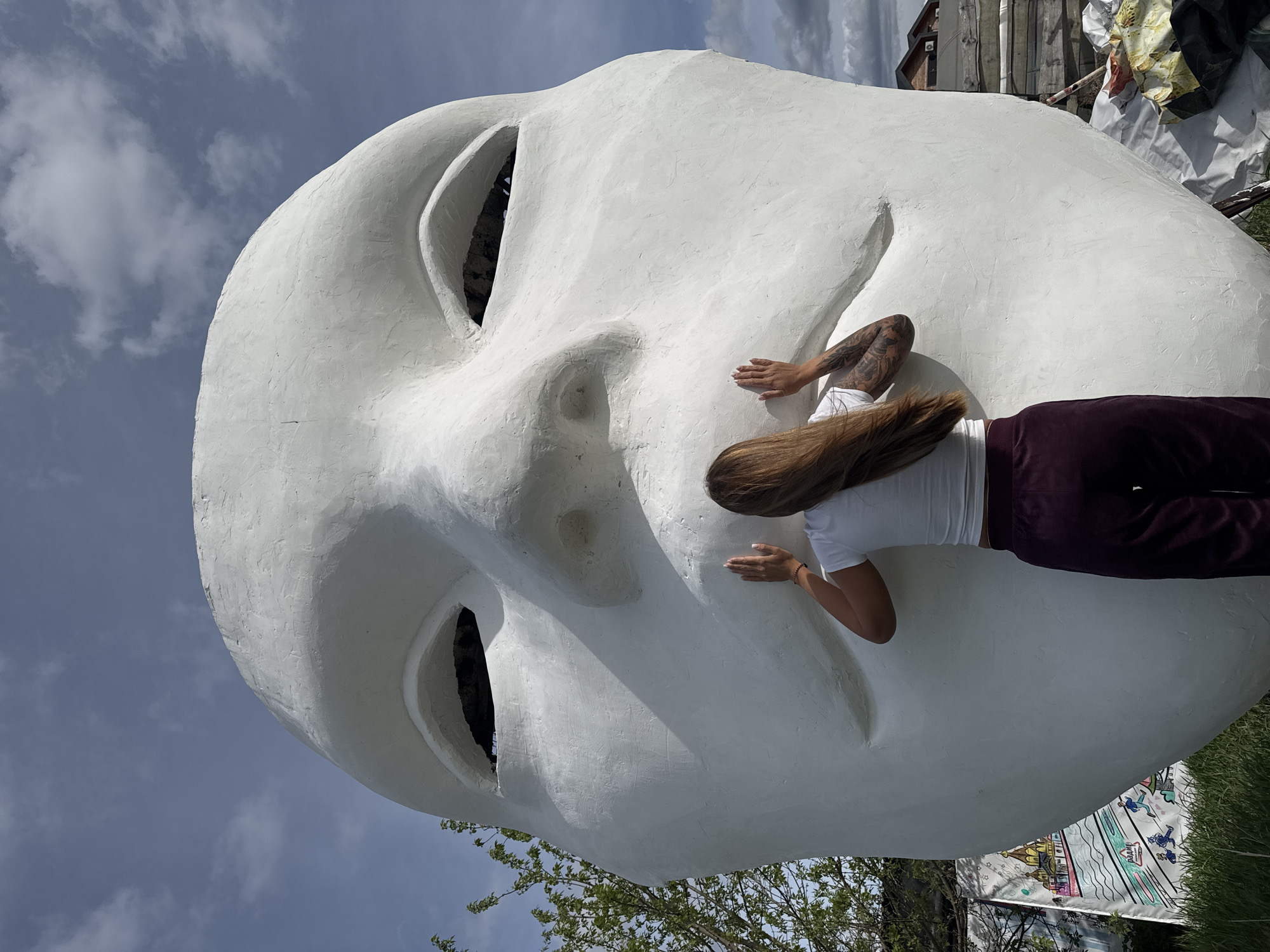 A woman touches the nose of a large white face sculpture located in Nizhny Novgorod, Russia. The face, featuring open eye sockets, dominates the scene against a backdrop of a partly cloudy sky. The woman is wearing a white shirt and dark burgundy-colored pants. Her hair is long and brown, covering the left side of her face as she reaches towards the nose of the sculpture. Tattoos are visible on her left arm. Behind the sculpture, there is some foliage, as well as a wooden structure. The picture is taken at a tilt.