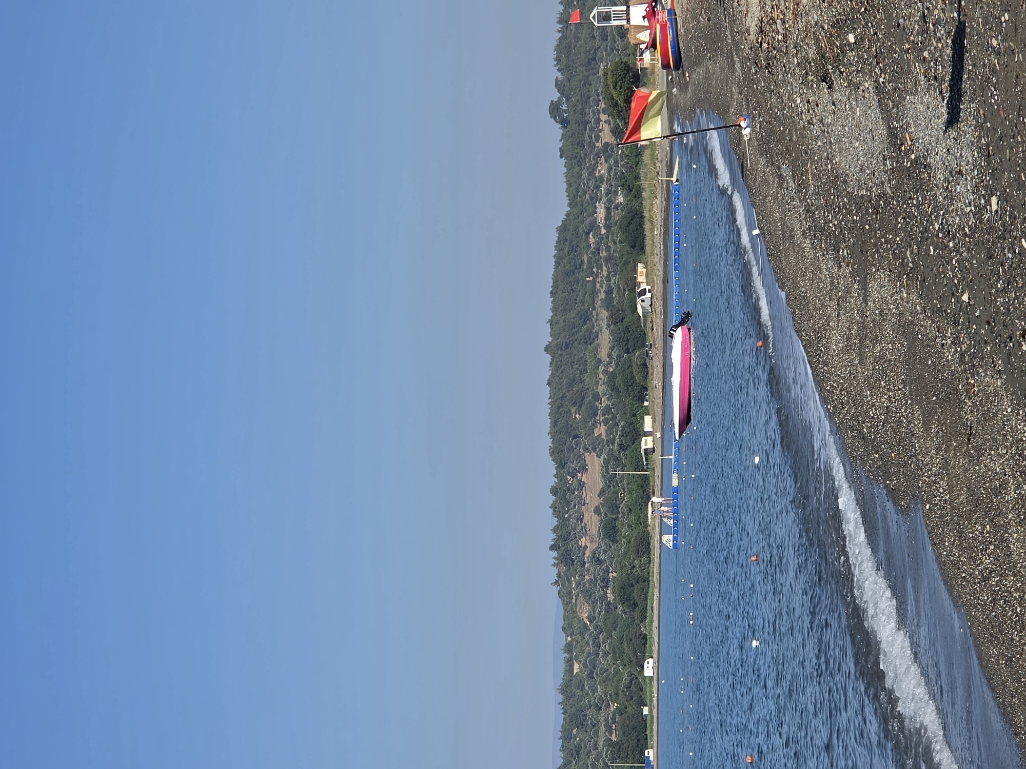 A daytime, eye-level, full outdoor shot shows a serene beach scene on a clear, sunny day. The sky is a vast expanse of uninterrupted blue. The tranquil sea, a deep blue, meets a dark, pebbled beach in the foreground, with gentle waves lapping at the shore.
In the water, a bright pink and white motorboat is visible. Further out, a blue floating platform with several people standing on it is present. Orange and white buoys dot the water, marking a boundary.
Along the shoreline, a lifeguard tower stands, adorned with a red flag at its peak. In front of the tower, a red and yellow striped flag waves on a pole. A red and blue inflatable boat is pulled up on the beach.
The background features a lush green hillside covered in trees and shrubs. The presence of some white camper vans or caravans suggests a campsite or recreational area along the coast. The general atmosphere is peaceful and inviting, indicative of a tourist destination. This scene is located in the town of Seferihisar, Turkey, known for its beautiful coastline.