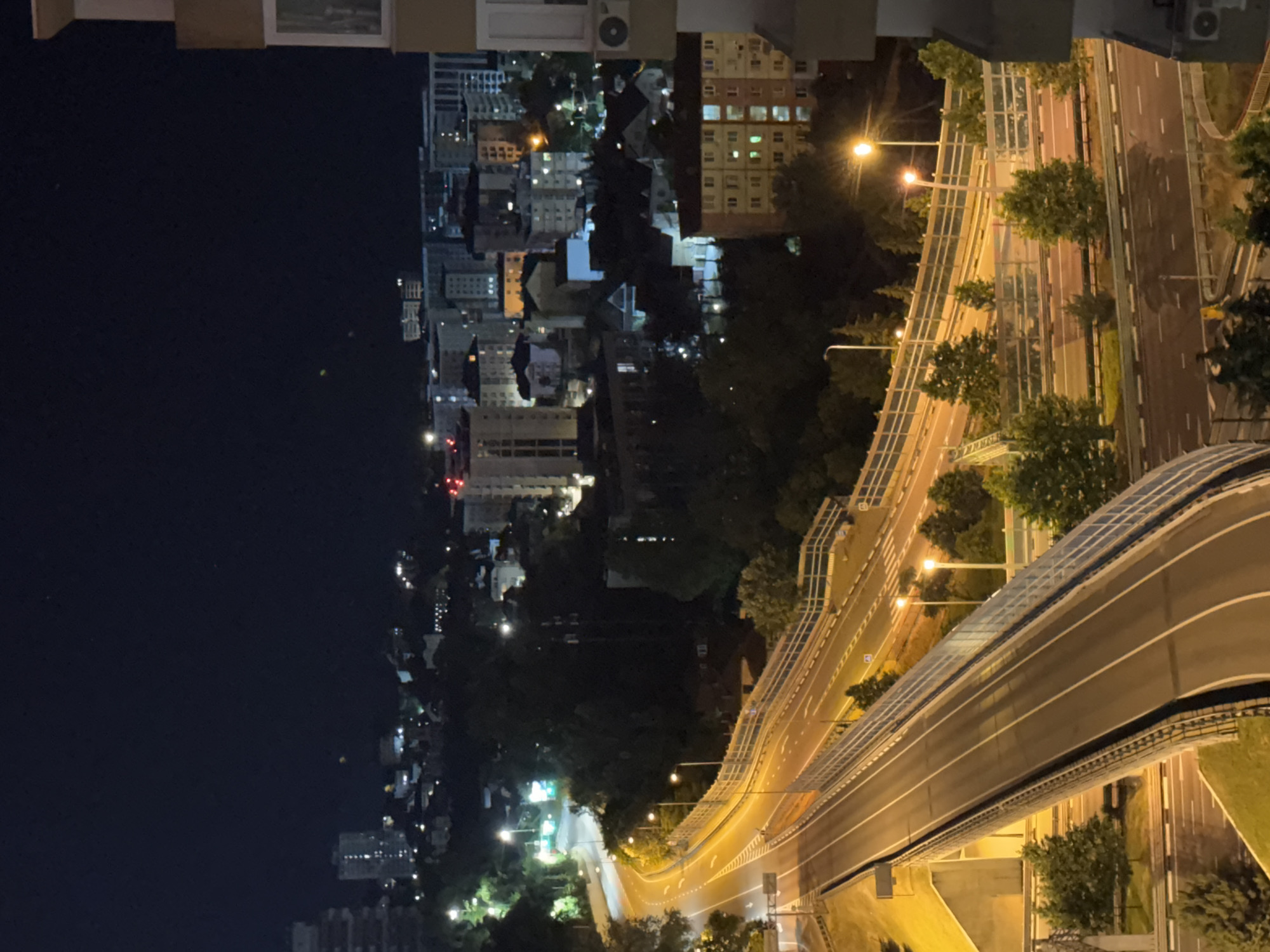 This is a nighttime aerial view of a complex road system in Sochi, Russia. The scene features multiple elevated roadways and bridges curving through a dark, tree-filled landscape. Streetlights illuminate the asphalt, casting long shadows and creating a warm glow on the roads and surrounding infrastructure. In the background, a city skyline is visible with numerous apartment buildings and structures, some of which are lit up, suggesting residential areas. The image captures a typical urban evening scene, with the infrastructure of transportation dominating the foreground and the city lights stretching into the distance.