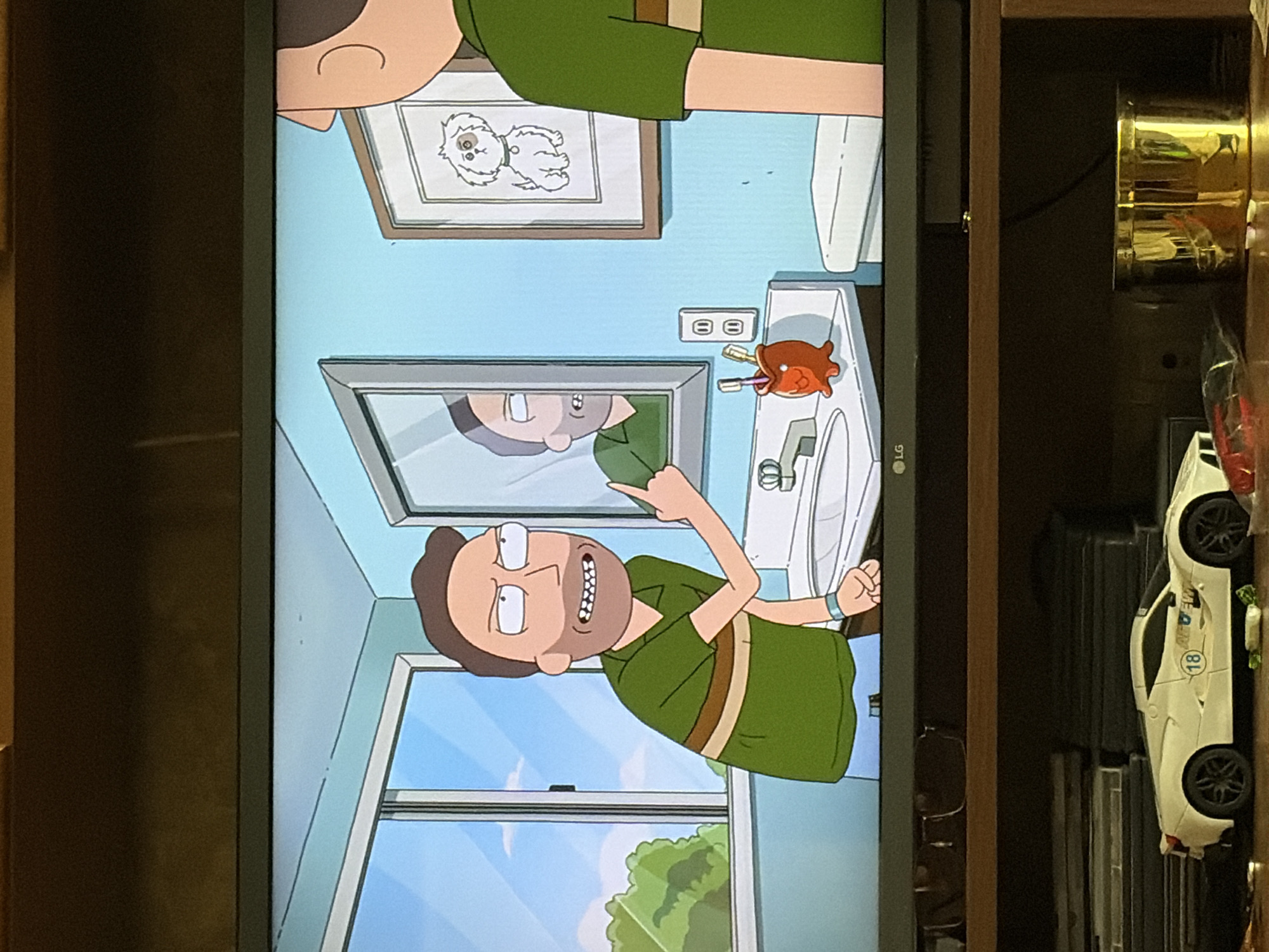 In a bathroom, a male animated character with brown hair and a beard stands at a sink, gesturing with his right hand. He wears a green shirt with brown stripes. A mirror reflects his image and another animated character with a beard. A framed picture of a white dog hangs on the light blue wall. A toothbrush holder shaped like an orange creature with a red face sits on the counter next to the sink. A white toilet is visible to the right. The scene is indoors, with a window showing greenery outside. Below the television, a white toy car with the number 18 on its side is present.