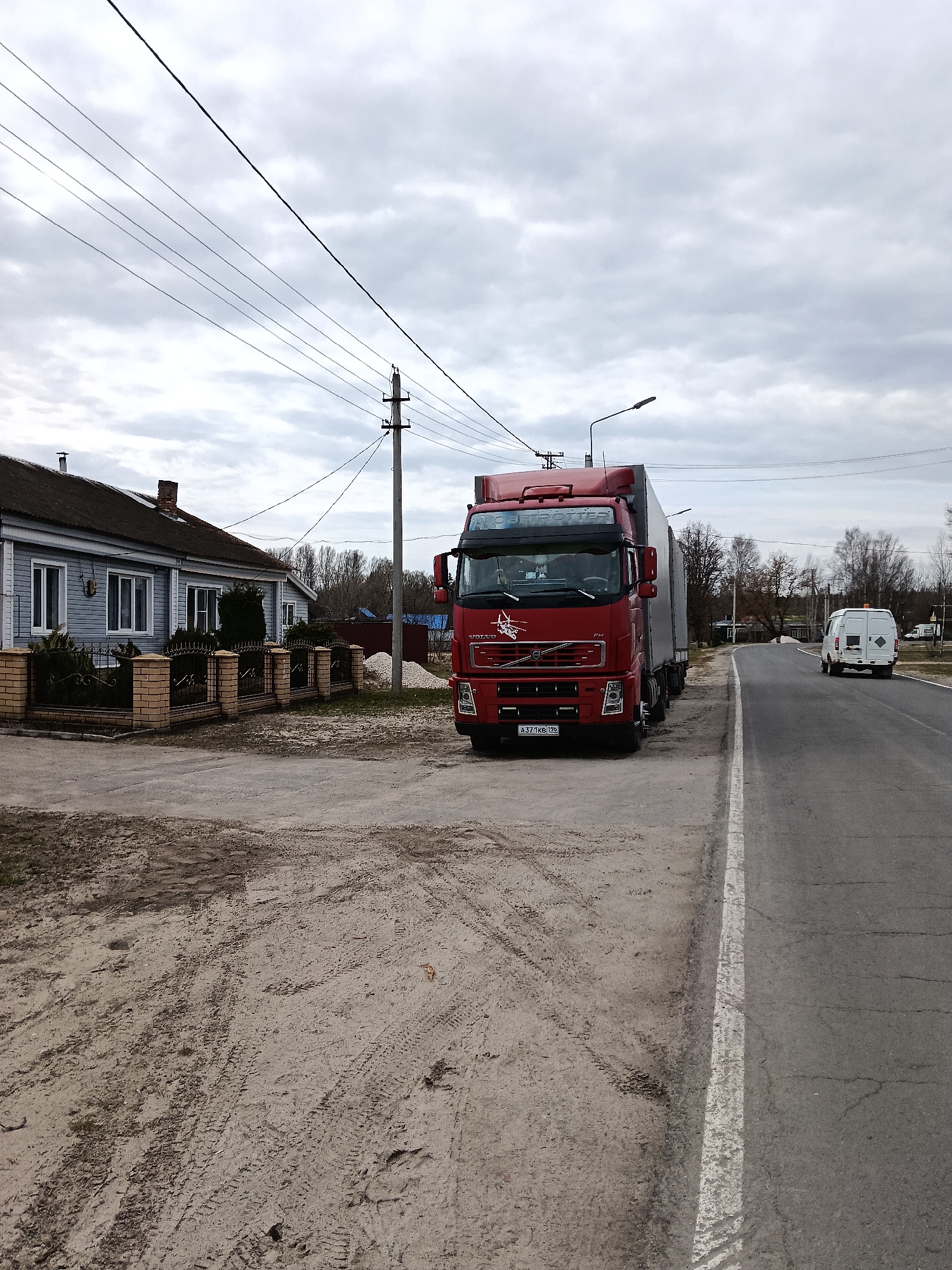 The image depicts a rural street scene in Красный Маяк, Russia, under an overcast sky. A red Volvo FH semi-truck is parked partially off the asphalt road on a dirt shoulder. A white van is visible driving on the road in the distance. To the left of the truck is a light blue, single-story house with white-framed windows and a brick and wrought-iron fence. Power lines and utility poles run along the side of the road. The foreground shows tire tracks and loose dirt on the shoulder of the road.