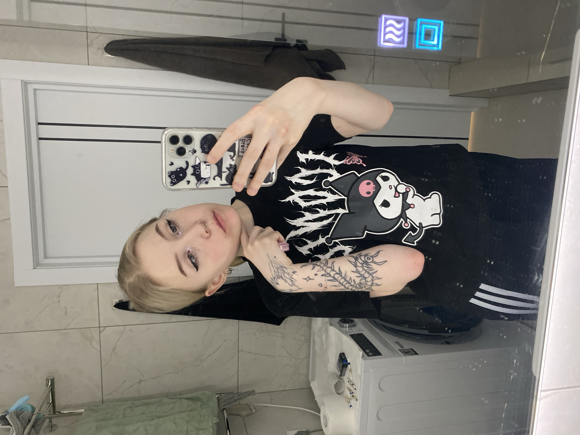 A selfie taken in a bathroom, with a woman as the main subject. She has blond hair, nose piercings, and eyeliner. She's holding a phone with a decorated case, wearing a black t-shirt with a cartoon print and what appears to be Adidas track pants. The background shows bathroom elements: a washing machine, tiled walls, a towel rack with a towel, a door, and a mirror with digital icons at the top. There is no evidence to suggest the image was taken in the city of Izhevsk, Russia.