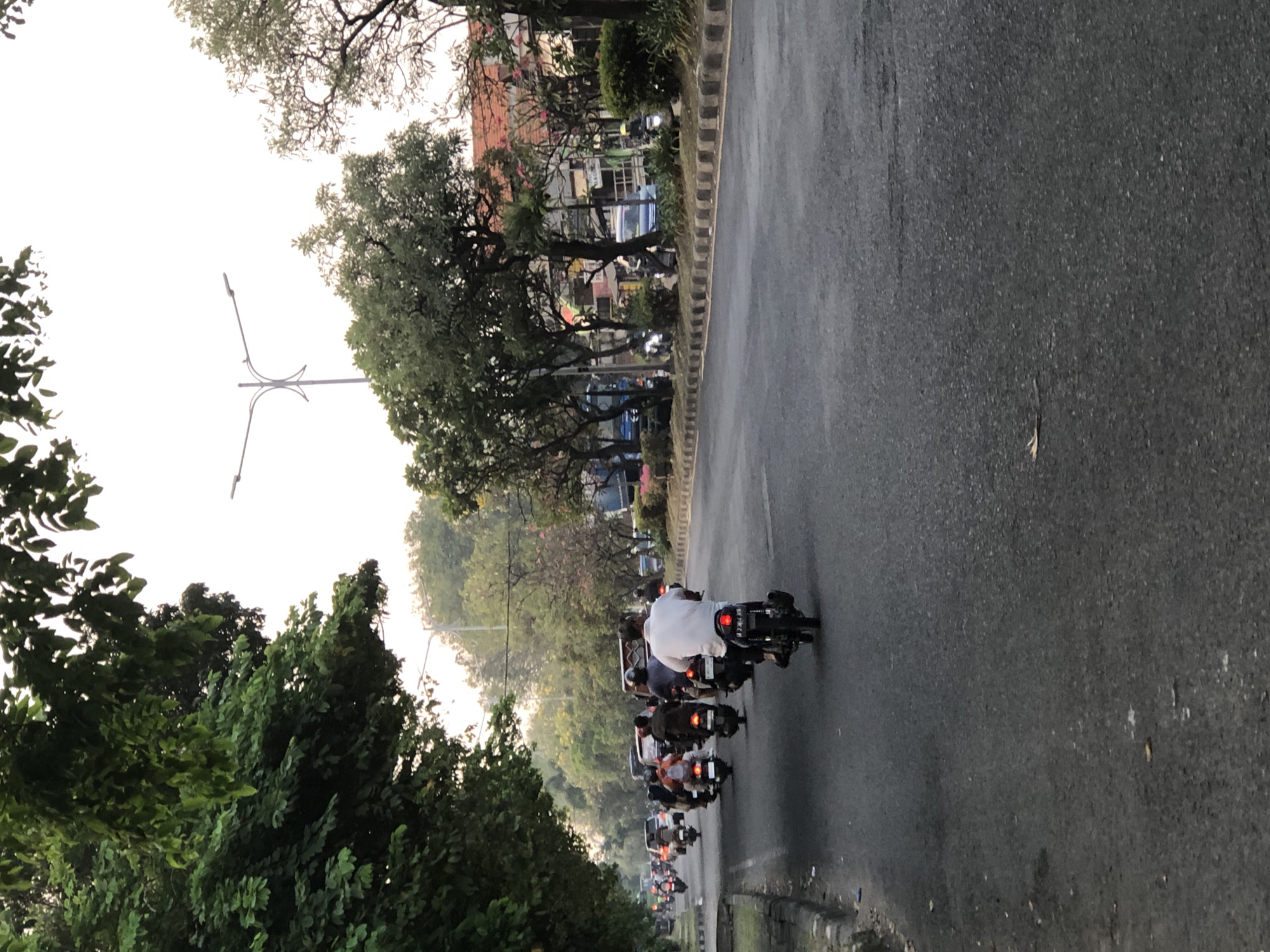 This is a photograph taken on a street in Sumorame, Indonesia. The image captures a traffic scene with numerous motorcycles and a few cars on an asphalt road. The lighting suggests it is either early morning or late afternoon, with the sky appearing overcast or hazy. Lush green trees line the left side of the road, with their branches extending over the pavement. On the right side of the road, there are buildings and more trees, with some flowers visible. Several individuals are seen riding motorcycles, some of whom are wearing white or colored shirts, and helmets. The vehicles are moving in the same direction, indicating a flow of traffic. The street has visible lane markings and a curb on the left side.