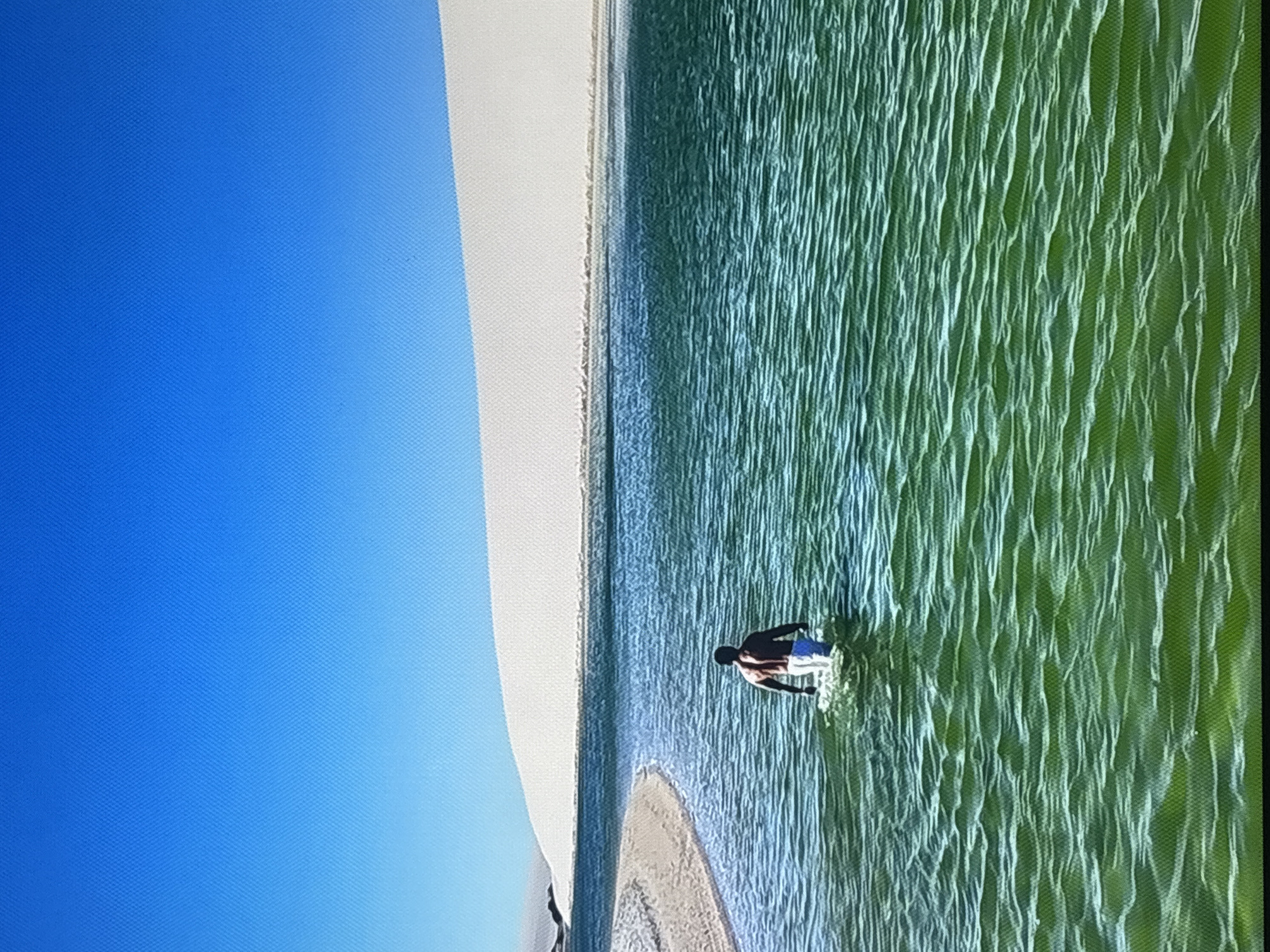 The image shows a person wading in a calm body of water with a bright blue sky overhead. To the left of the water is a large, light-colored sand dune that slopes upwards. The water itself has a greenish hue and is rippling, suggesting a gentle breeze. The person is in the middle ground, facing away from the camera and towards the sand dune. They are wearing dark shorts and a striped top. The scene appears to be tranquil and serene. The bright sky suggests it is daytime, likely sunny. While the location is provided as Valladolid, Spain, there are no specific visual cues within the image itself to confirm this. The image does not contain any visible text.