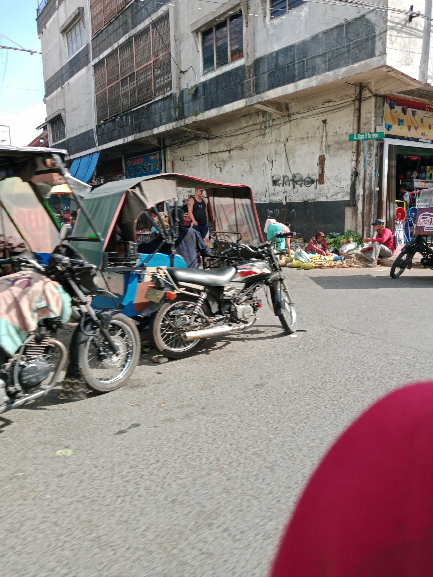 A street scene in the City of Binjai, Indonesia, shows a street with several motorbikes and a trishaw parked. A building with a weathered facade stands on the left, featuring multiple windows with barred coverings and faded paint. A street sign reads "Jl. Kapt. P. Tea Dean." In the background, people are visible tending to a market stall with produce laid out on the ground, including bananas and other vegetables. The ground is paved with asphalt.