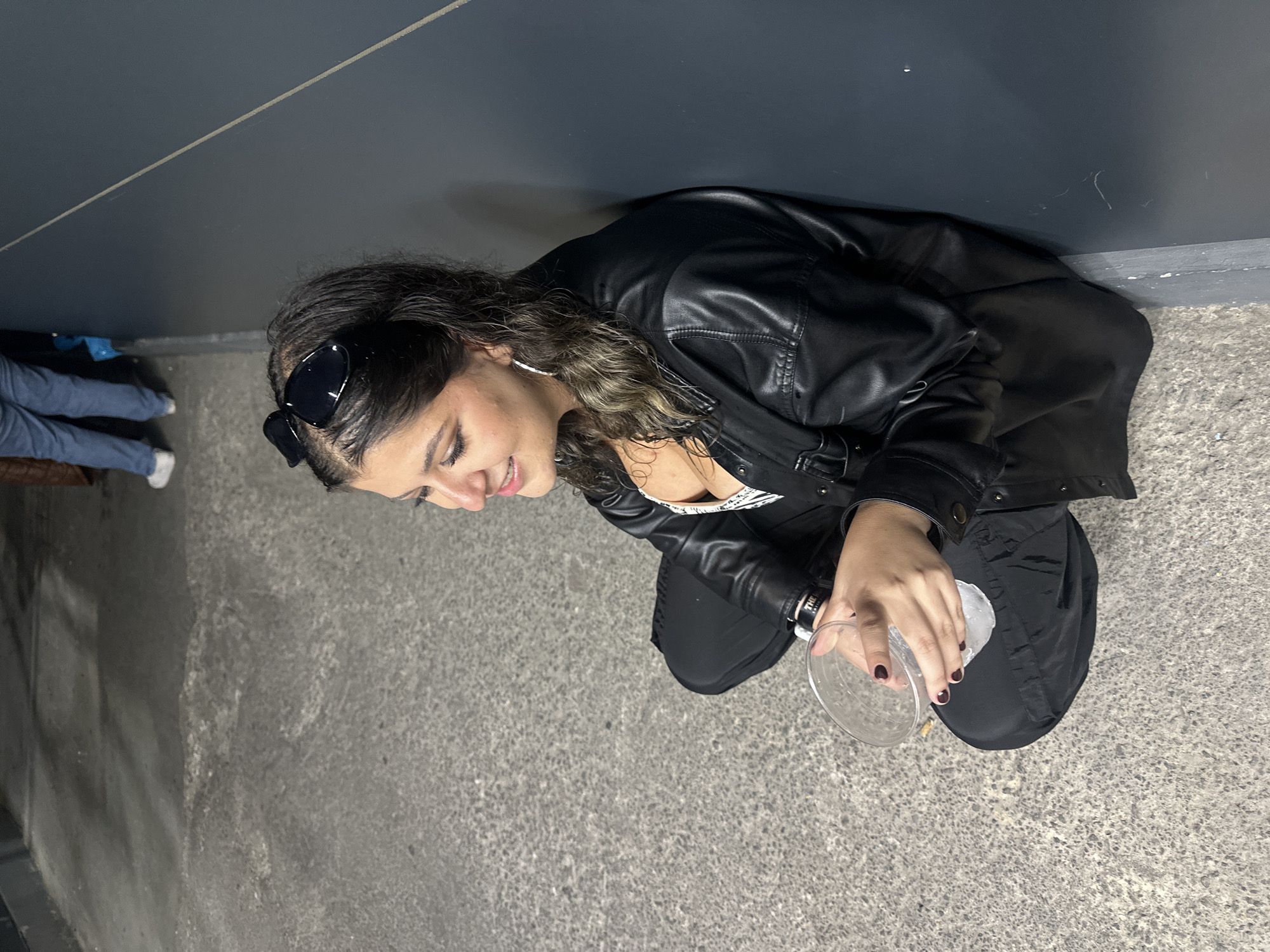 A woman with long, wavy hair, wearing sunglasses and a black leather jacket, squats on a concrete surface. She is holding a clear plastic cup with ice in her right hand. A wall is behind her. Other people are partially visible in the background. The scene appears to be in Mexico City, Mexico.
