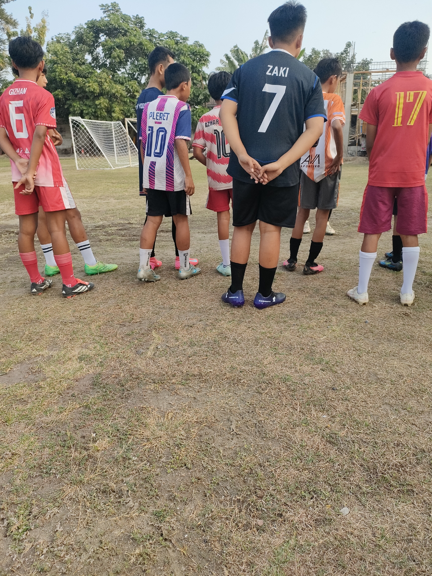 The image depicts a group of young boys, likely a soccer team, standing on a dry, grassy field with patches of dirt. In the background, a soccer goal with a white net is visible, along with trees and some construction materials. The boys are wearing soccer uniforms and cleats.
Specifically, the scene is located at a soccer field in the Banguntapan sub-district, Bantul Regency, Yogyakarta, Indonesia. One boy in the foreground wears a red jersey with the number "6" and the name "GIZHAN" on the back. To his right, another boy is in a blue and pink striped jersey with "PLERET" and "10". To the right of him, a boy in a red and white striped jersey has "AZHAR" and "10" on his back. A boy in the center wears a dark blue jersey with "ZAKI" and "7" on the back, and black shorts. To his right, a boy is in an orange and white striped jersey. On the far right, a boy wears a red jersey with "17" on the back and dark red shorts.
The ground is covered with dry, sparse grass and sandy soil. The lighting suggests it is daytime, likely late afternoon, with the sun casting soft shadows. The overall atmosphere is one of preparation or a break during a soccer practice or match.