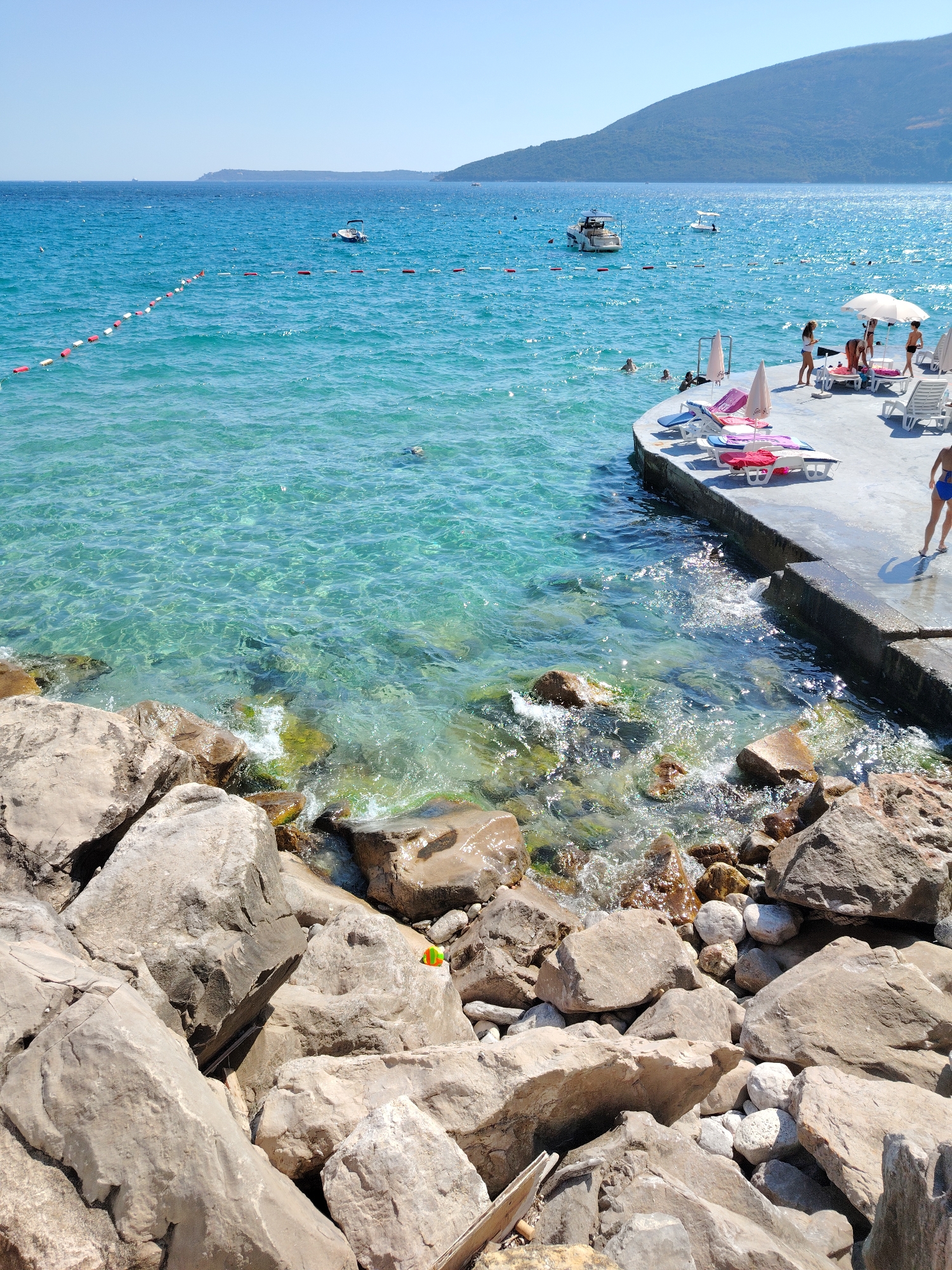 The image depicts a sunny day at a rocky beach and a concrete pier in Herceg Novi, Montenegro. The crystal-clear turquoise water laps against the shore, revealing the rocky seabed. A string of red and white buoys marks a swimming area in the sea. Several people are visible, some swimming in the water, while others are relaxing on lounge chairs on the pier. A white umbrella provides shade on the pier, where beach towels and loungers are arranged. Two boats are anchored in the distance, and a lush green hillside forms the backdrop. The foreground is dominated by large, weathered rocks and smaller pebbles.
