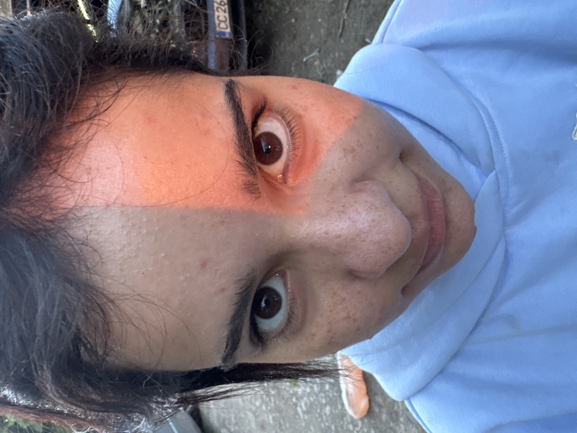 A close-up, angled portrait of a woman with dark hair, brown eyes, and freckles. She is wearing a light blue hoodie. Part of her face is illuminated with an orange hue. Behind her are indistinct shapes and colors, including parts of a vehicle license plate with "CC 26" visible. The background suggests an outdoor location, but the image does not provide enough information to pinpoint the location in Ruzhitsa, Bulgaria.