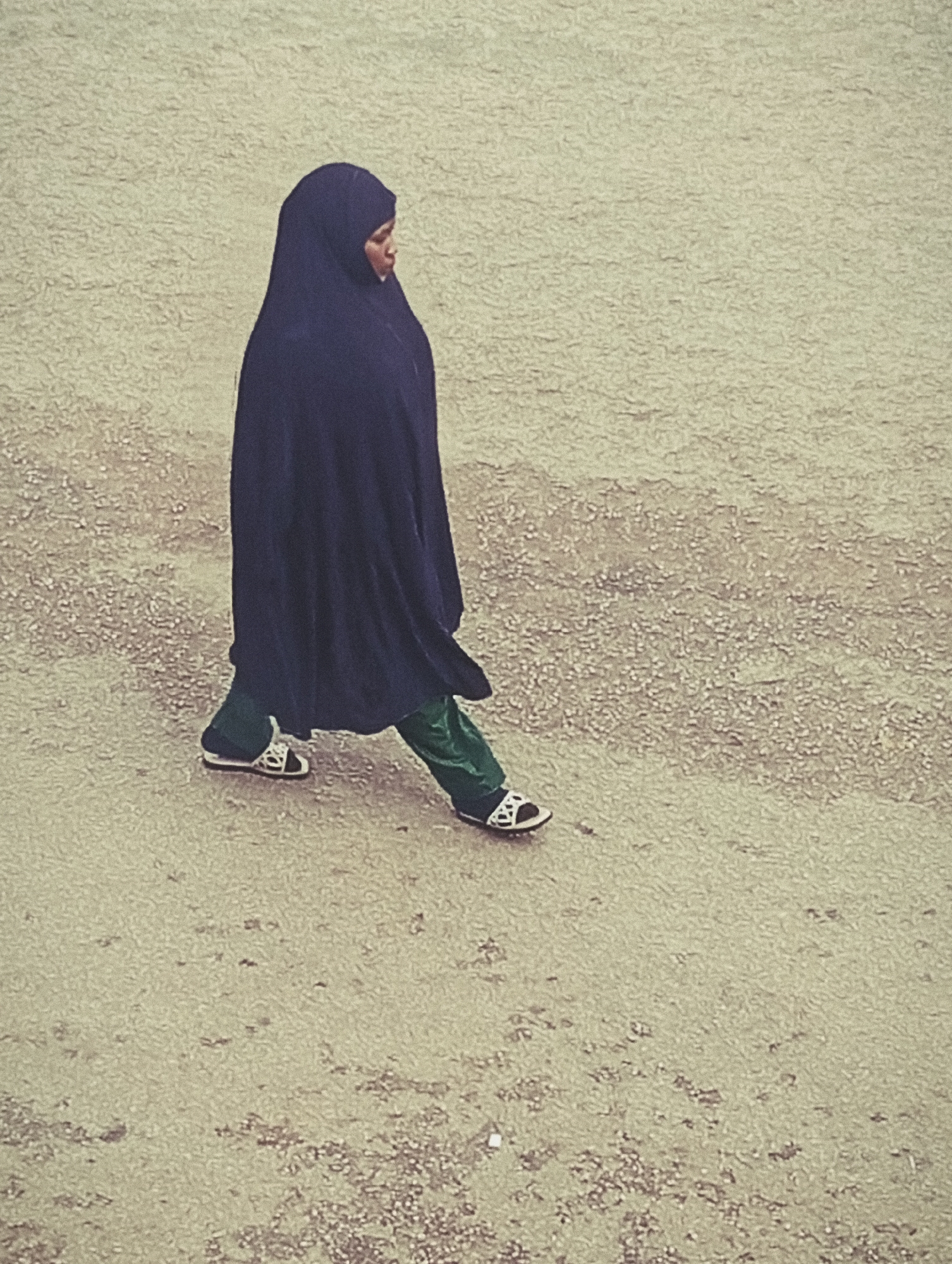 A full-body shot of a woman walking across a textured, sandy surface. The woman is wearing a dark navy headscarf that drapes over her shoulders, obscuring most of her body. Her legs are clad in bright green trousers that are visible beneath the hem of her headscarf. She is wearing white sandals with black straps and a black sole. Her left leg is forward, mid-stride, while her right leg is behind her. The woman is looking to her right, with her head turned slightly away from the camera.
The surface she is walking on appears to be a mixture of sand and small pebbles or grit, with a few scattered larger stones. The lighting suggests it is daytime, but the overcast sky creates a diffused light, with no strong shadows. The overall impression is of a muted, perhaps slightly hazy atmosphere. The image is composed from a high-angle perspective, looking down on the woman as she walks. The background is a continuation of the same sandy surface, with a subtle texture. There is no visible text in the image. The setting could be a pathway, a dusty yard, or an unpaved area in a rural or semi-urban location, consistent with a location in Bukuru, Nigeria.