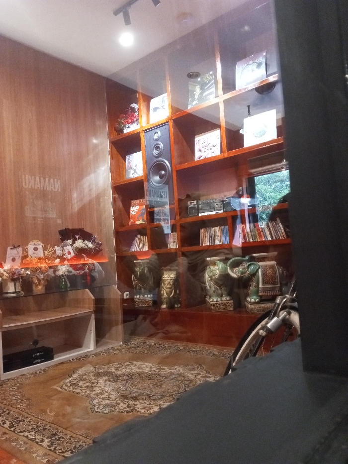 The scene depicts the interior of a commercial or public establishment in Pontianak, Indonesia, observed through a transparent glass partition.
To the left, a light-toned wooden paneled wall features a reversed reflection of text resembling "NAMAKU". Against this wall, a multi-tiered glass display unit holds various decorative items, including multiple small floral arrangements in vases and other wrapped objects. A dark electronic console, consistent with a gaming system, occupies a lower, opaque shelf.
The central and right portions of the interior are dominated by a dark brown wooden shelving unit with numerous open compartments. These shelves contain an assortment of books, compact discs, or vinyl records. A large, black audio speaker with a prominent central woofer is vertically positioned within one of the upper shelf compartments. Below the speaker, a reversed reflection displaying text, likely "TENAMAAST", is visible on the glass. A silver boombox-style stereo system with dual speakers rests on a higher shelf. On the lower shelves and the floor, at least three large, intricately patterned ceramic elephant statues are prominently displayed. A window in the upper right background reveals blurred green foliage from an outdoor environment.
The floor within the establishment is covered by a large, patterned oriental-style rug. Recessed spotlights are visible on the ceiling, providing interior illumination. The immediate foreground consists of a dark, unidentifiable opaque surface. In the lower right foreground, outside the glass partition, the front wheel and a portion of the frame of a bicycle are visible. No individuals are present within the frame.