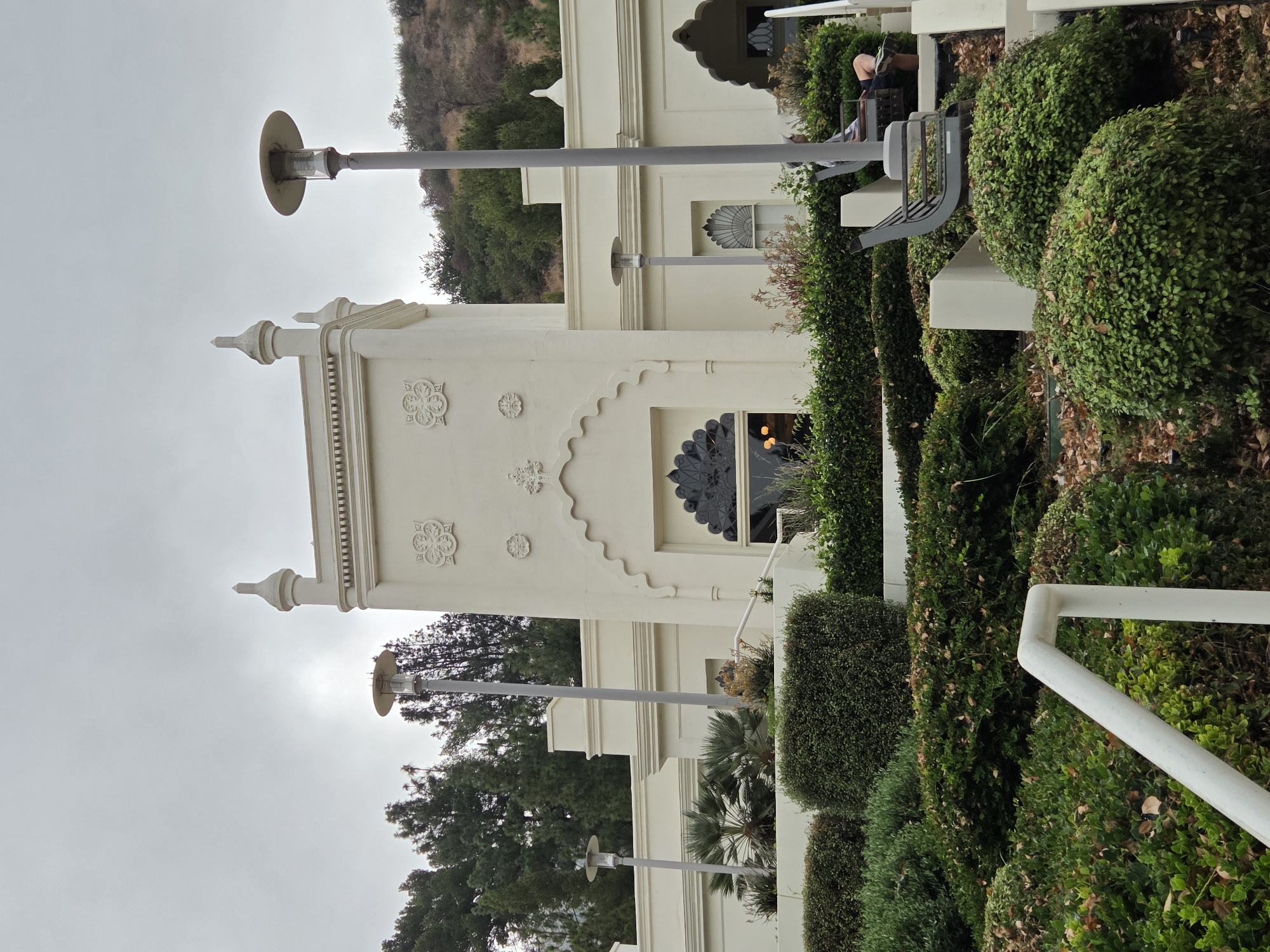 The image shows the exterior of the Self-Realization Fellowship Lake Shrine in Glendale, California, United States of America. The building is white with ornate architectural details, including arched doorways and windows with decorative grilles. A central tower is topped with finials. The building is surrounded by lush green landscaping, including manicured bushes and trees. Two tall, modern lampposts are visible in front of the building. A person is seated on a bench on the right side of the image, amidst the greenery. The sky is overcast.
