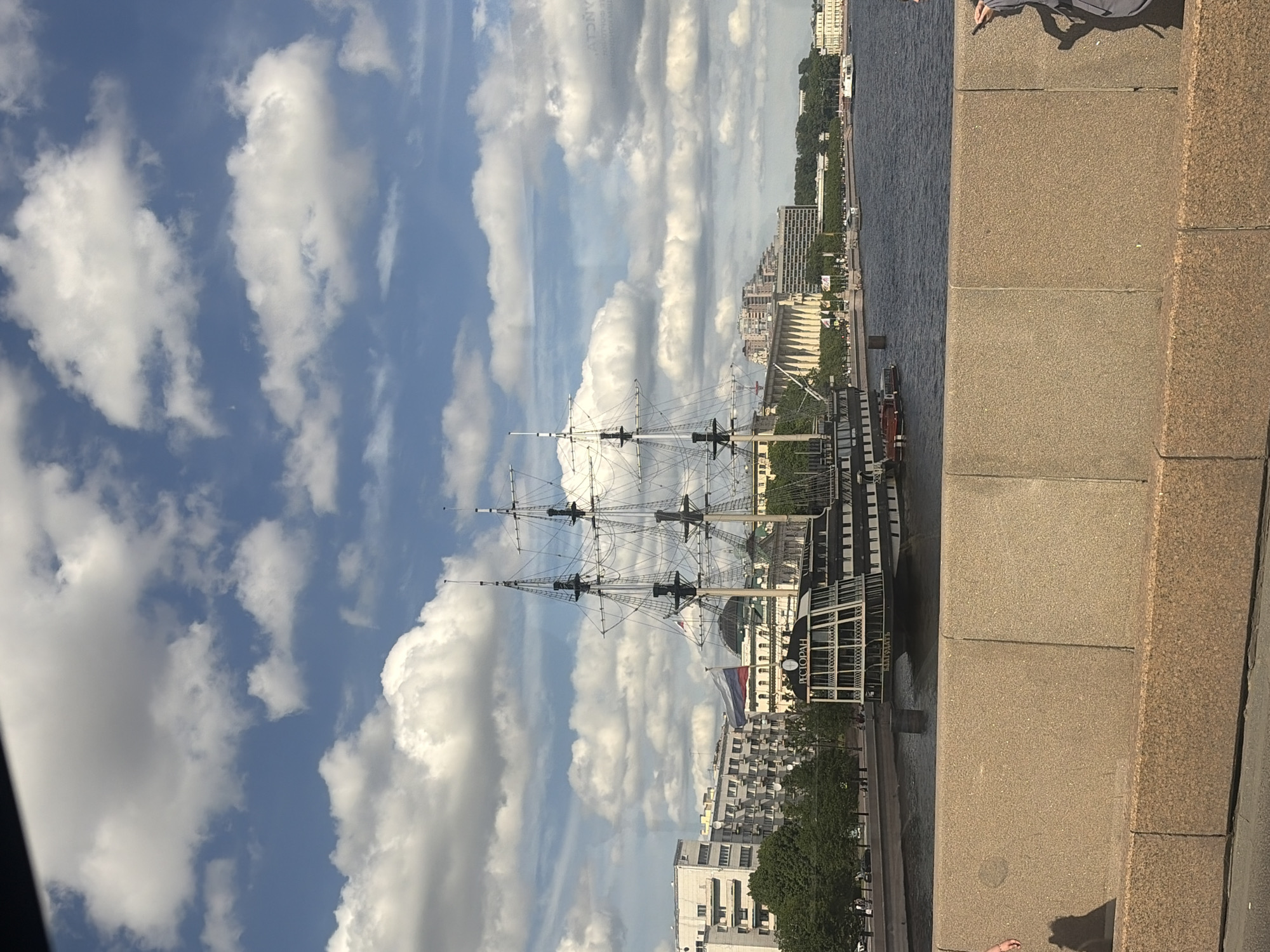 The image captures a view of the Neva River in Saint Petersburg, Russia, as seen from a bridge. Dominating the foreground is a stone embankment. In the middle ground, a large, historic sailing ship, possibly a museum or restaurant, is moored near the embankment. The ship features multiple masts and rigging. Behind the ship, the cityscape of Saint Petersburg is visible, with several buildings, including residential blocks and classical architecture, lining the riverbank. Lush green trees are also present along the waterfront. The sky above is a vibrant blue, punctuated by numerous fluffy white clouds. The water of the Neva River appears dark and reflects the sky and surrounding structures. A Russian flag is prominently displayed on the ship.