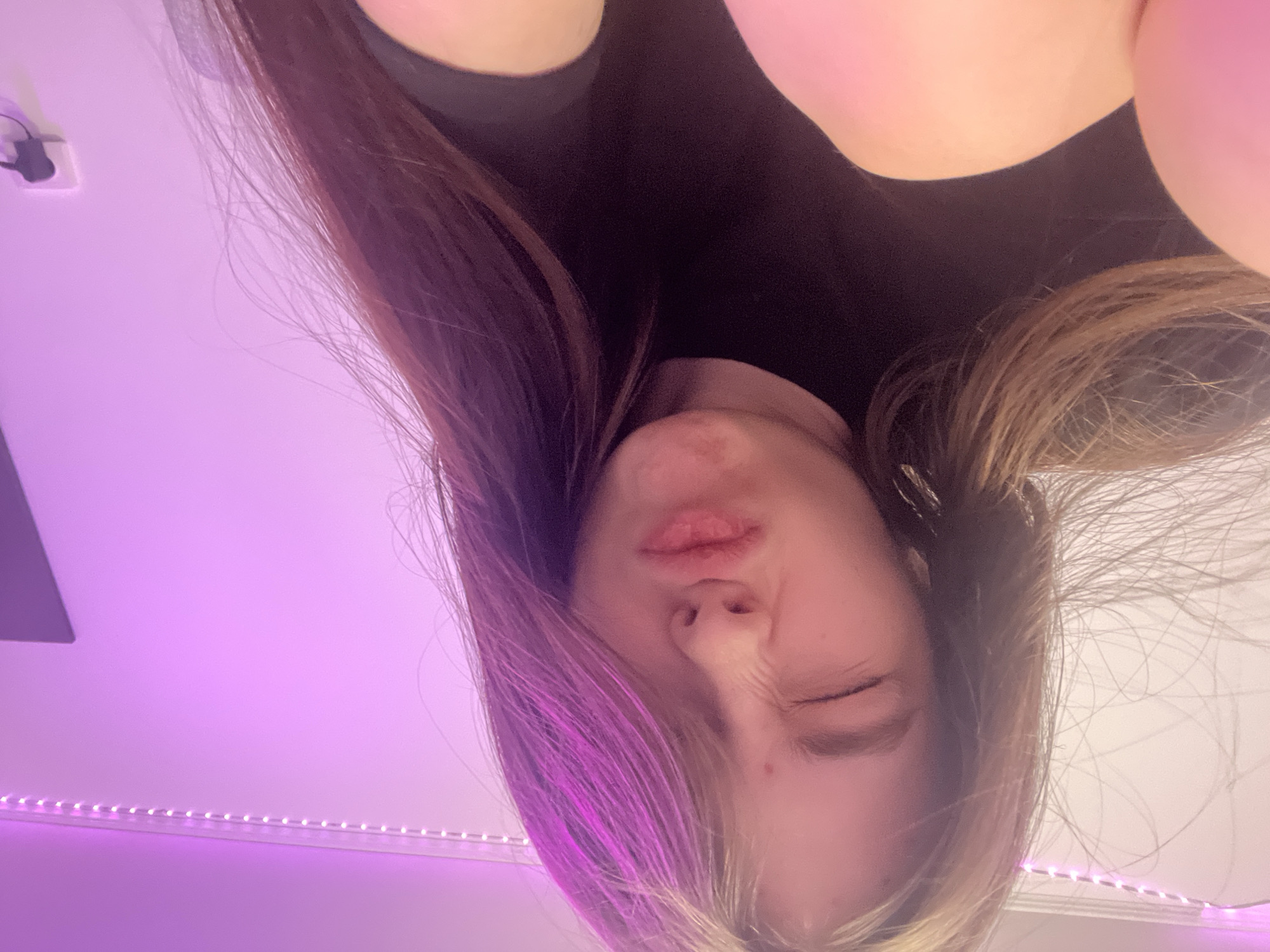 A selfie taken from an inverted angle shows a person with long brown hair wearing a black top. One eye is closed, and the lips are puckered. The background consists of a white ceiling with pink LED strip lights near the edge. A white electrical outlet and a gray square object are visible on the ceiling. The location is not discernible from the image.