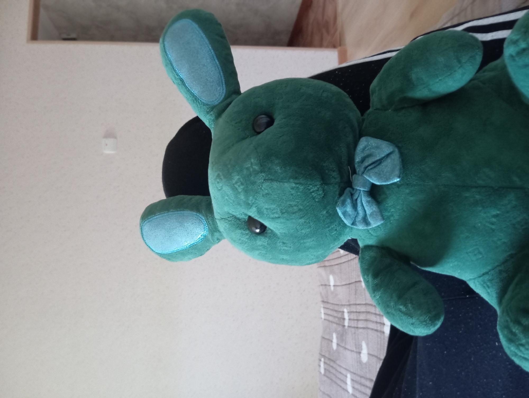 A plush green bunny toy with blue ear inserts and a blue bowtie is seated against a black surface, possibly a chair or lap. The bunny has round black eyes and appears to be facing forward. Behind the bunny, a wall with a textured pattern is visible, along with a white light switch. To the left, a bed with a grey and white striped blanket is partially visible. To the right, a doorway or opening leads to another room with wooden flooring and a white door frame. The lighting suggests an indoor environment during the day. There is no identifiable location in the city of Temryuk, Russia.