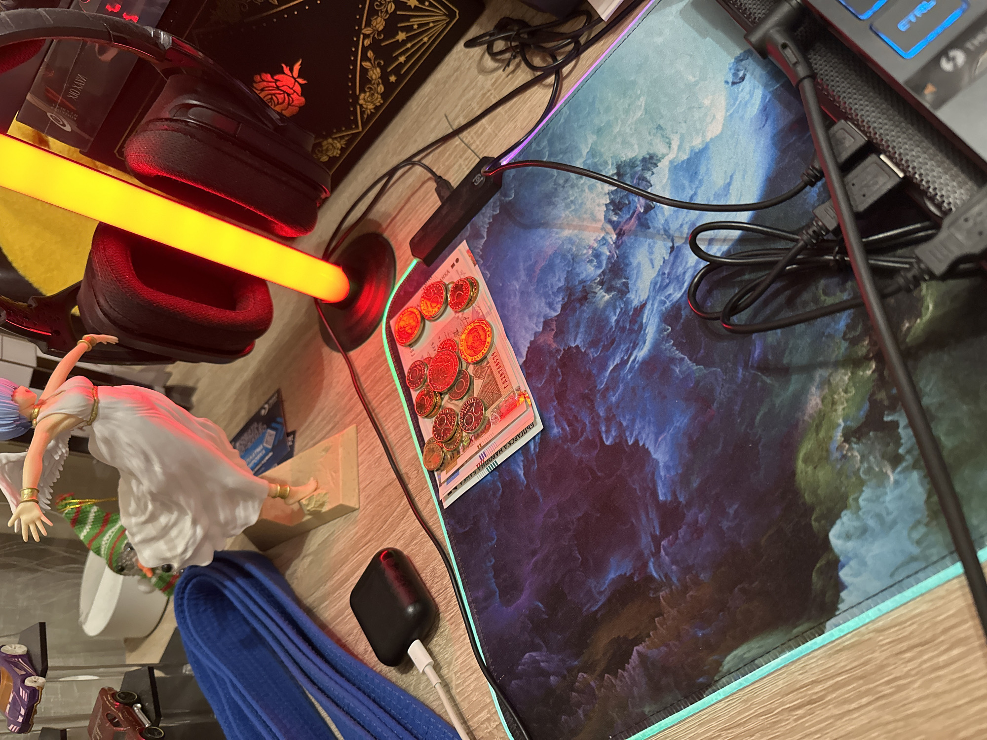 The scene depicts a detailed personal desk setup located in an interior space in the city of Unknown, Unknown. The primary surface is a light-toned wooden desk, likely laminate. The lighting is predominantly artificial, featuring a prominent vertical yellow/orange LED lamp on the right side and an RGB LED strip outlining a gaming mouse pad, displaying teal and purple hues.
In the upper-left corner, two miniature vehicles are displayed on clear acrylic risers: a purple and gold sports car on top, and a maroon/red hot rod below. To the left, a blue fabric item, resembling a rolled martial arts belt, rests on the desk.
Centrally positioned on the desk is a large gaming mouse pad featuring a nebula or cosmic landscape design with vibrant blues, purples, and greens. On the mouse pad, a Bulgarian 100 leva banknote is partially visible, oriented horizontally, with several small, reddish-gold coins (possibly Bulgarian stotinki) arranged on top of it and to its right.
A black Apple AirPods charging case, connected to a white USB-C cable, is actively charging on the mouse pad, slightly to the left of the banknote.
To the right, a figurine of a female character with light blue hair, a flowing white dress, and white wings stands on a beige base. She is depicted in an active pose with outstretched arms and gold bands on her wrists and ankles. Behind the figurine, a black gaming headset with red illuminated ear cups rests against the bright yellow LED lamp. Behind the headset and lamp, a dark, rectangular object, possibly a book or box, is visible with intricate gold detailing, including a rose motif and a fan-like design with stars.
In the lower-right corner, a cluster of black cables, including a black USB hub, is present. Part of a dark keyboard with backlit keys, showing a blue "CTRL" key, is visible at the extreme bottom-right. No people are visible in the image.