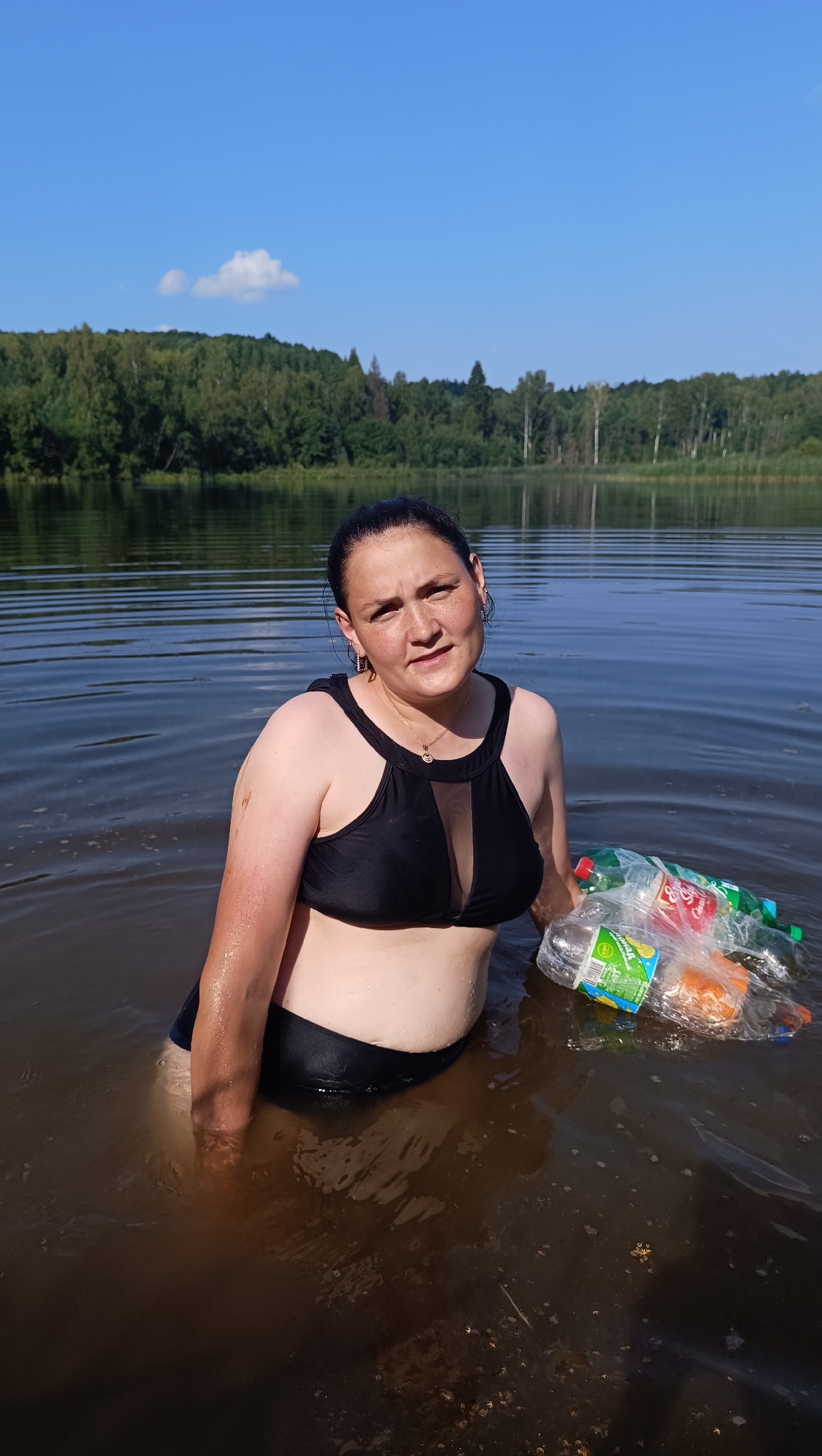 A woman is standing in a lake, wearing a black bikini. She is holding a bag filled with plastic bottles. The lake is surrounded by trees, and the sky is blue with some clouds. The water is murky. The scene appears to be in Karsha, Russia.