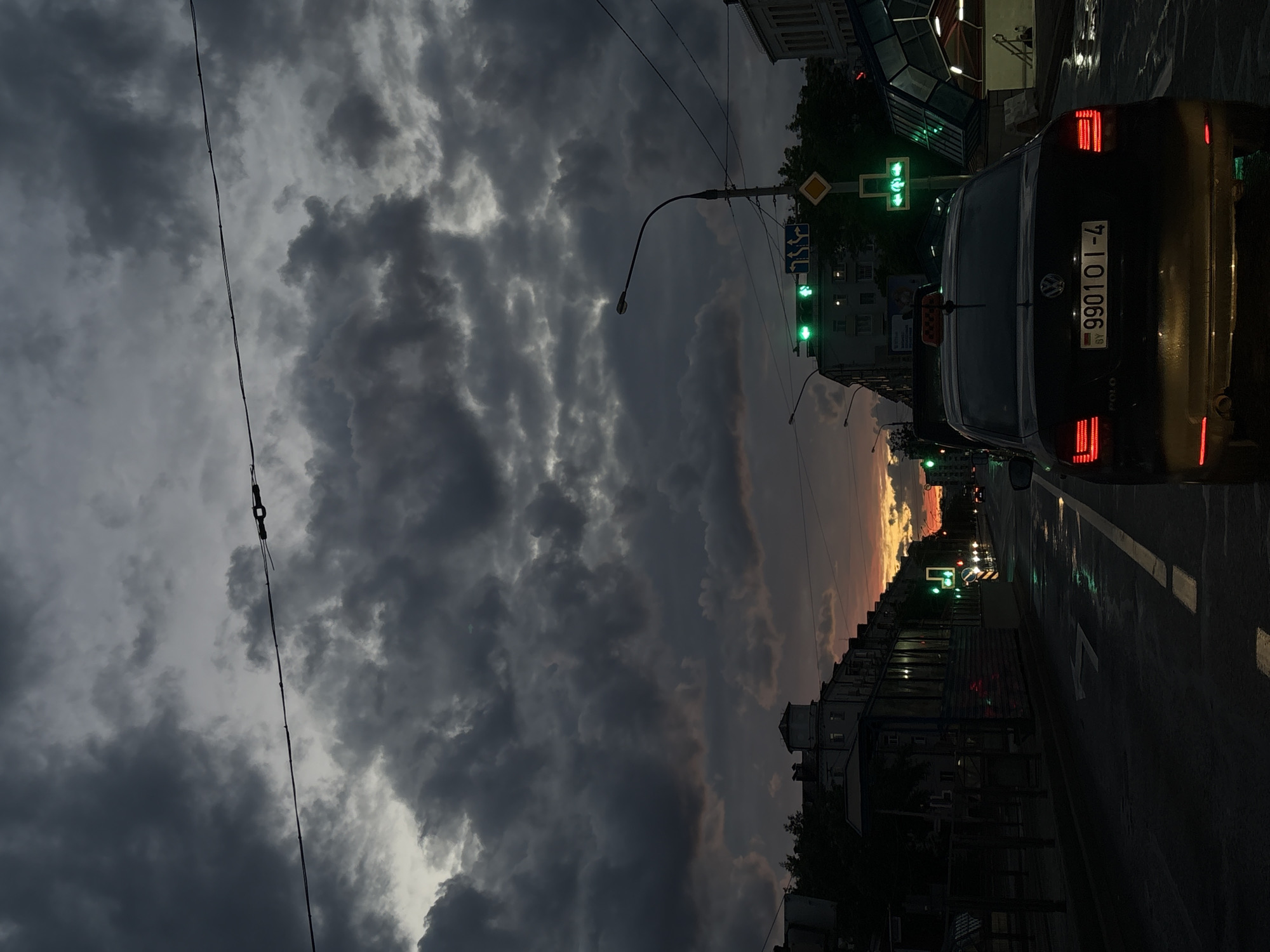 The image shows a street scene in Minsk, Belarus, at dusk. The sky is filled with heavy, dark storm clouds. A black Volkswagen Polo is in the right foreground, with a taxi indicator on top. The license plate number is 9901 OI-4 and is Belorussian. A street light and traffic light is on the right side of the street, showing green lights. A pedestrian crossing is visible with green lights as well. In the distance, the sunset casts an orange glow between buildings.