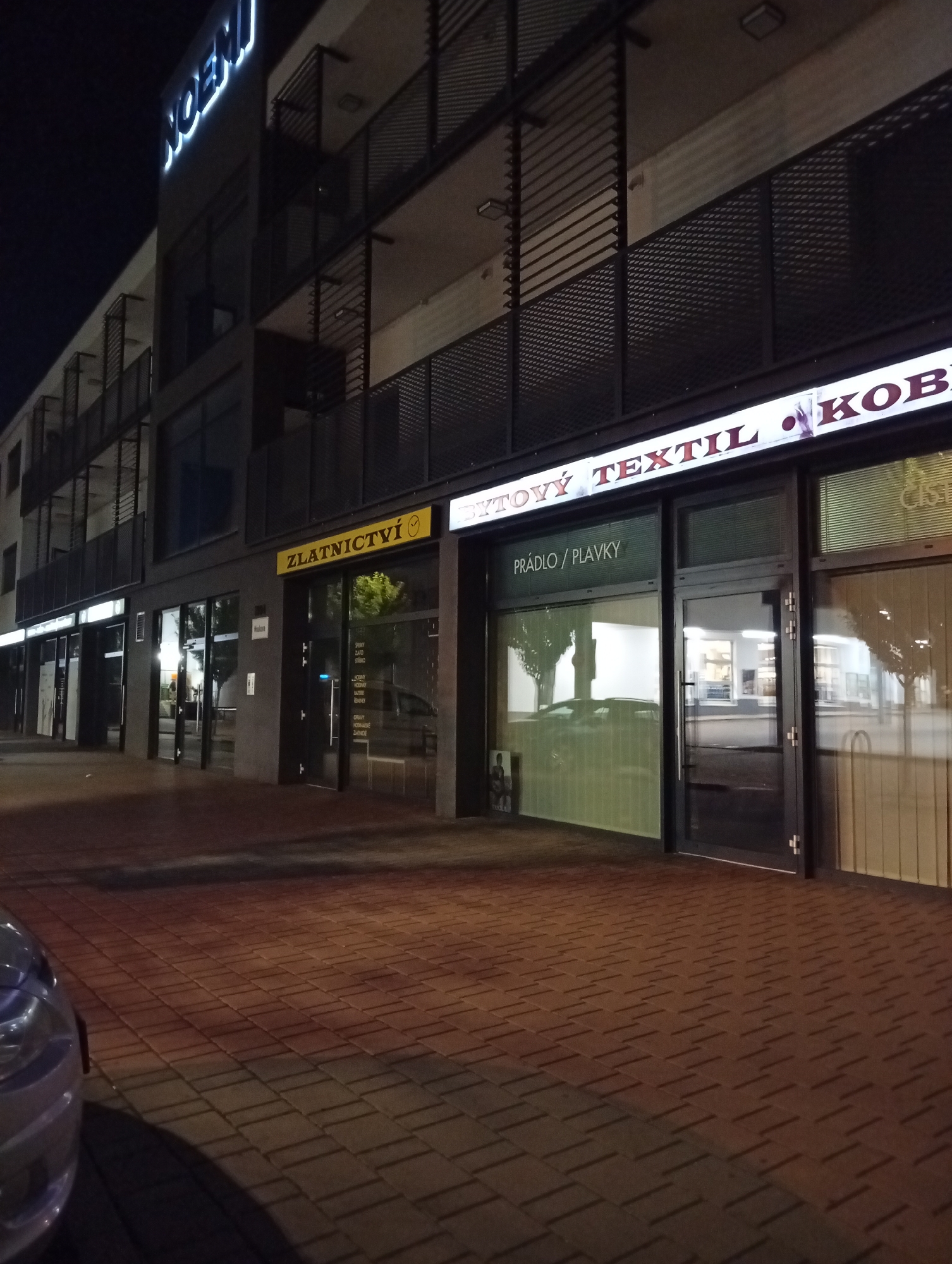 The image shows a nighttime exterior view of a row of shops in Tišnov, Czechia. The building has multiple stories with balconies. The shops have illuminated signs. From left to right, the signs read "NOEN", "ZLATNICTVÍ" (which translates to "Jewelry"), and "TEXTIL KOB". The shop labeled "TEXTIL KOB" also has a sign above its entrance reading "PRÁDLO / PLAVKY" (which translates to "Lingerie / Swimwear"). The ground is paved with brick-like material. A car is partially visible in the foreground on the left. The lighting suggests it is after dark, with streetlights and internal shop lighting illuminating the scene.