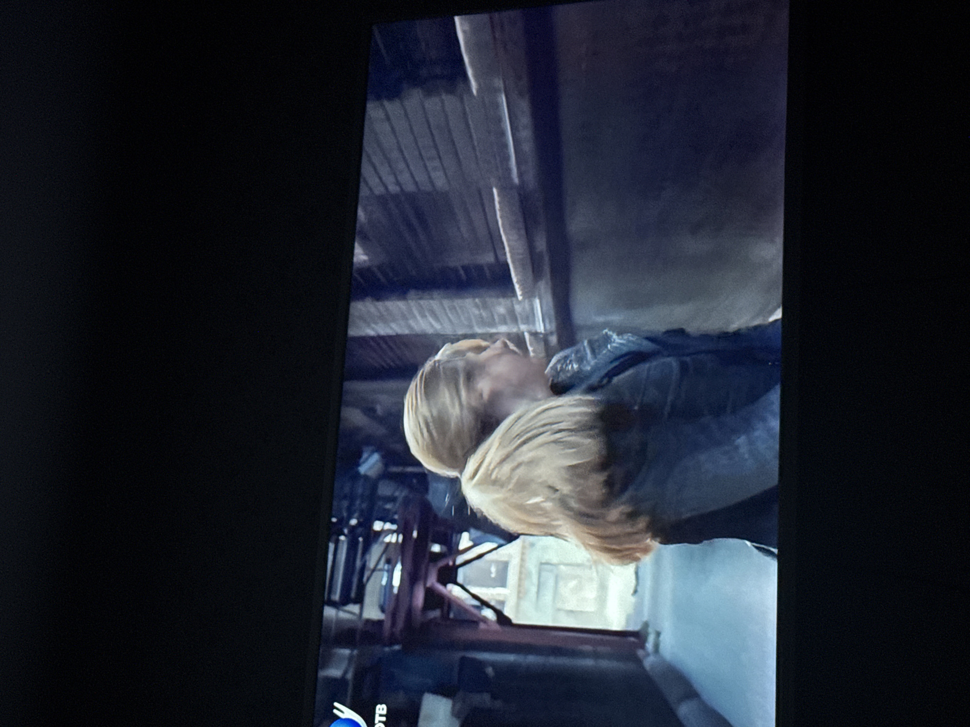 A scene depicted on a television screen shows a fair-skinned person with blonde hair in an urban environment, potentially Mytishchi, Russia. The setting features industrial elements, including metal structures and a wooden ceiling, with a weathered aesthetic. The person is wearing a dark-colored jacket. The image appears to be taken from a screen recording.