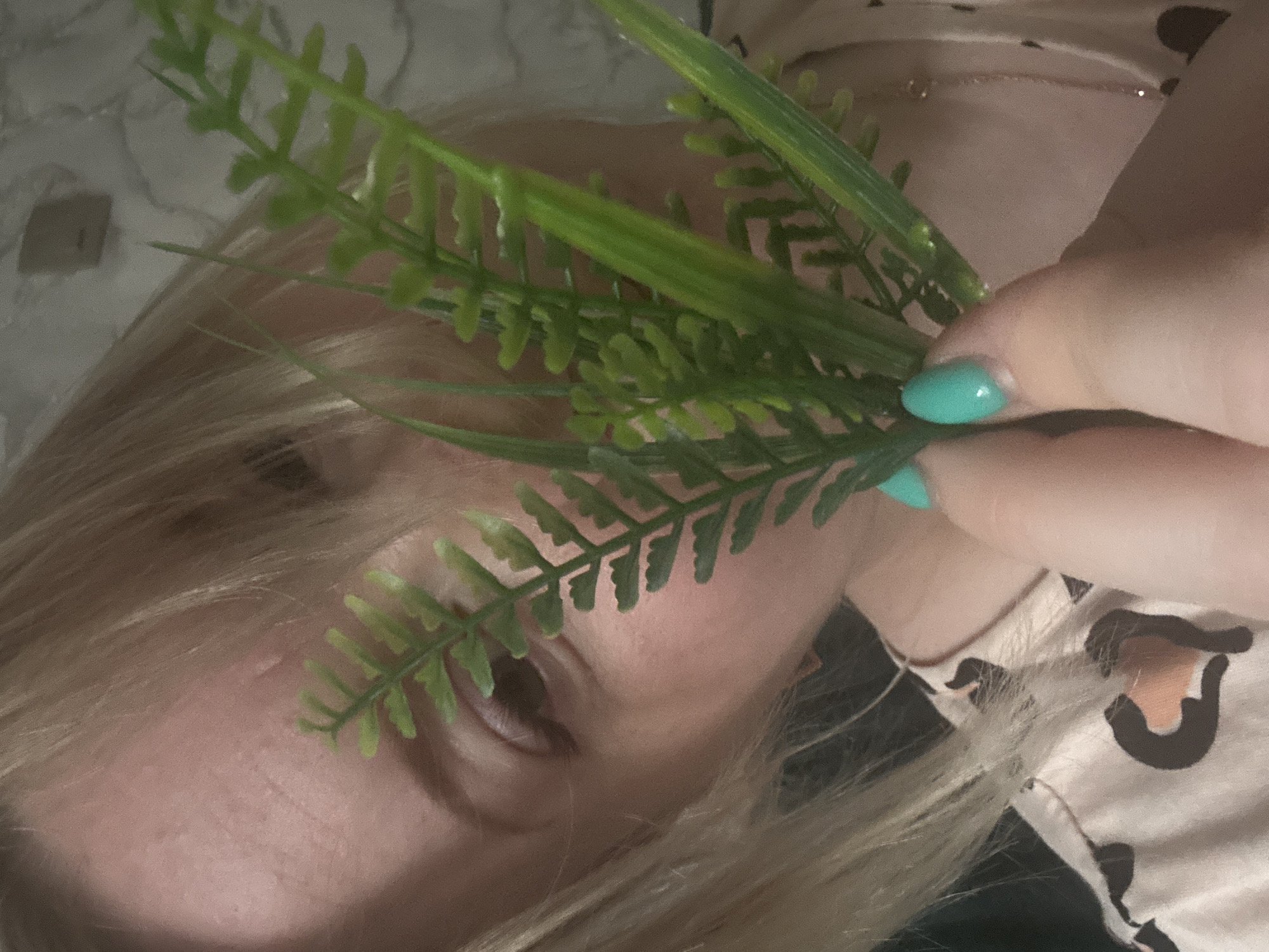 A close-up, slightly rotated image shows a woman with light blonde hair. Her left eye is visible through a handful of green artificial fern leaves held up by her right hand. She has turquoise nail polish on her nails. She is wearing a light-colored shirt with a dark animal print. The background appears to be a textured wall or surface. The location is unknown, and there is no indication that the image was taken in the city of Chaikovsky, Russia.