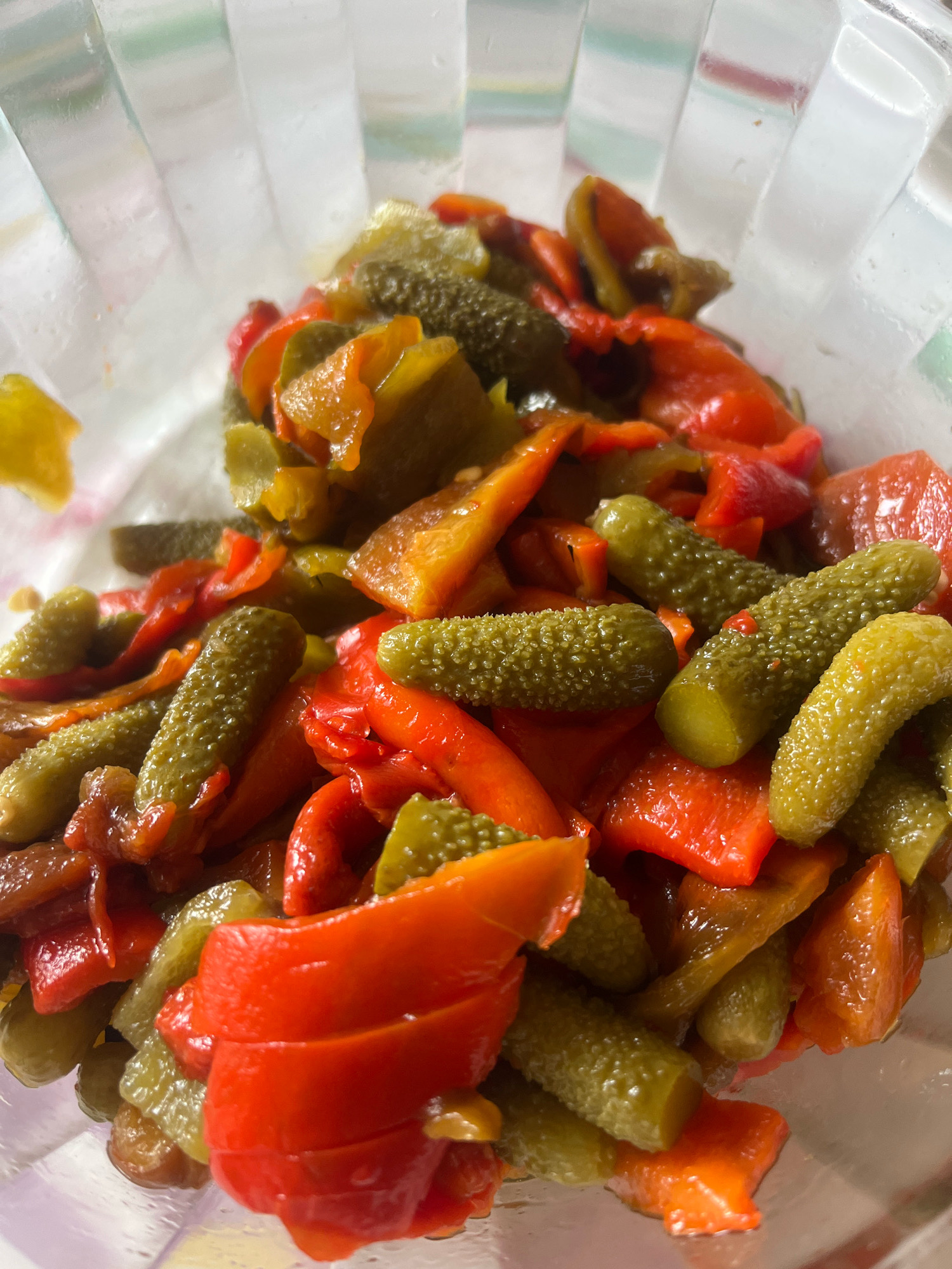 A close-up, overhead shot shows a glass bowl filled with a mixture of chopped red bell peppers and small, bumpy gherkins. The peppers appear to be roasted or pickled, with some showing slightly caramelized edges. The gherkins are a vibrant green and whole. The bowl is clear and has vertical ridges on the outside. The background is out of focus and appears to be a light-colored surface, possibly a table. There are no people or animals visible in the image. The scene is indoors, likely a kitchen or dining area in Cheshnegirovo, Bulgaria, given the context.