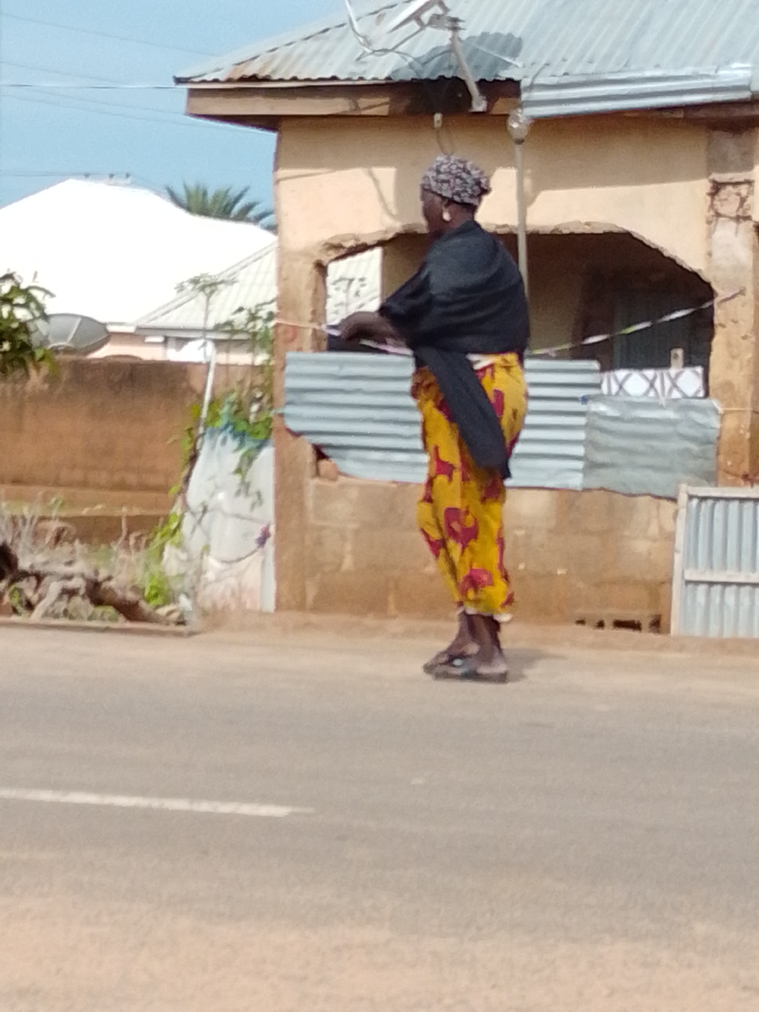 In Gombe, Nigeria, a person wearing a black top and a yellow and red patterned skirt stands facing away from the camera on a dusty road. The person is wearing a headscarf and earrings, and appears to be holding something in their hands. In the background, a building with a corrugated metal roof and a light brown wall is visible. To the left of the building, a white structure with a satellite dish on top is partially obscured by greenery. The road has a white line marking its center.