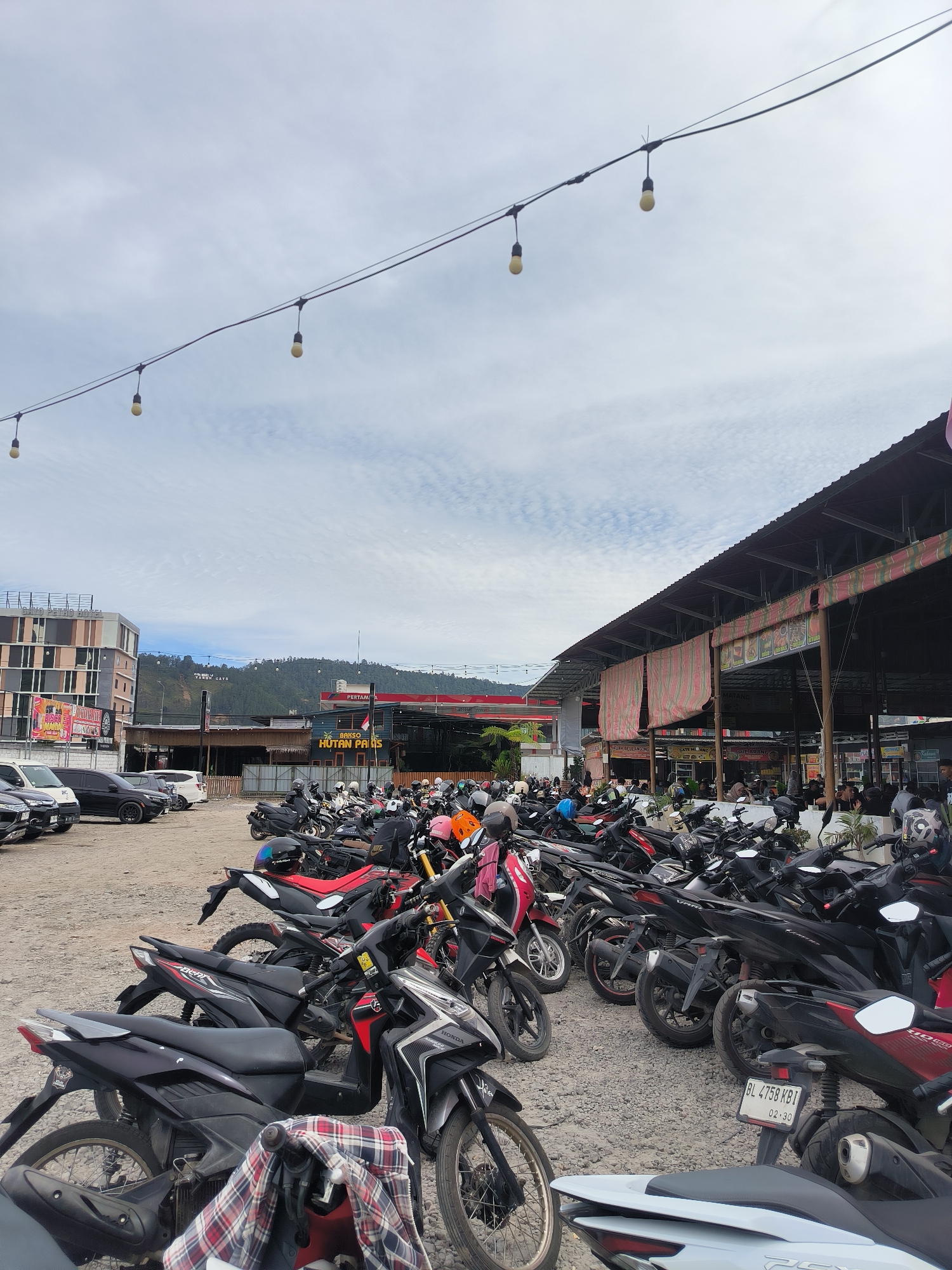 The image depicts a crowded outdoor parking lot filled with numerous motorcycles in Nunangantara, Indonesia. The parking lot is adjacent to a covered outdoor dining area with multiple food stalls, indicated by signage such as "BAKSO HUTAN PAIS" and "KUAH BIEBANGE" and other unreadable text. String lights are suspended overhead, and a multi-story building, possibly a hotel with signage "BUKIT RETRO HOTEL," is visible in the background, along with a hillside featuring a large sign with white lettering. The ground is covered with gravel. Several people are seen sitting at tables within the dining area, and others are standing or walking around the stalls. The sky is partly cloudy. The scene appears to be a busy commercial area during daylight hours.