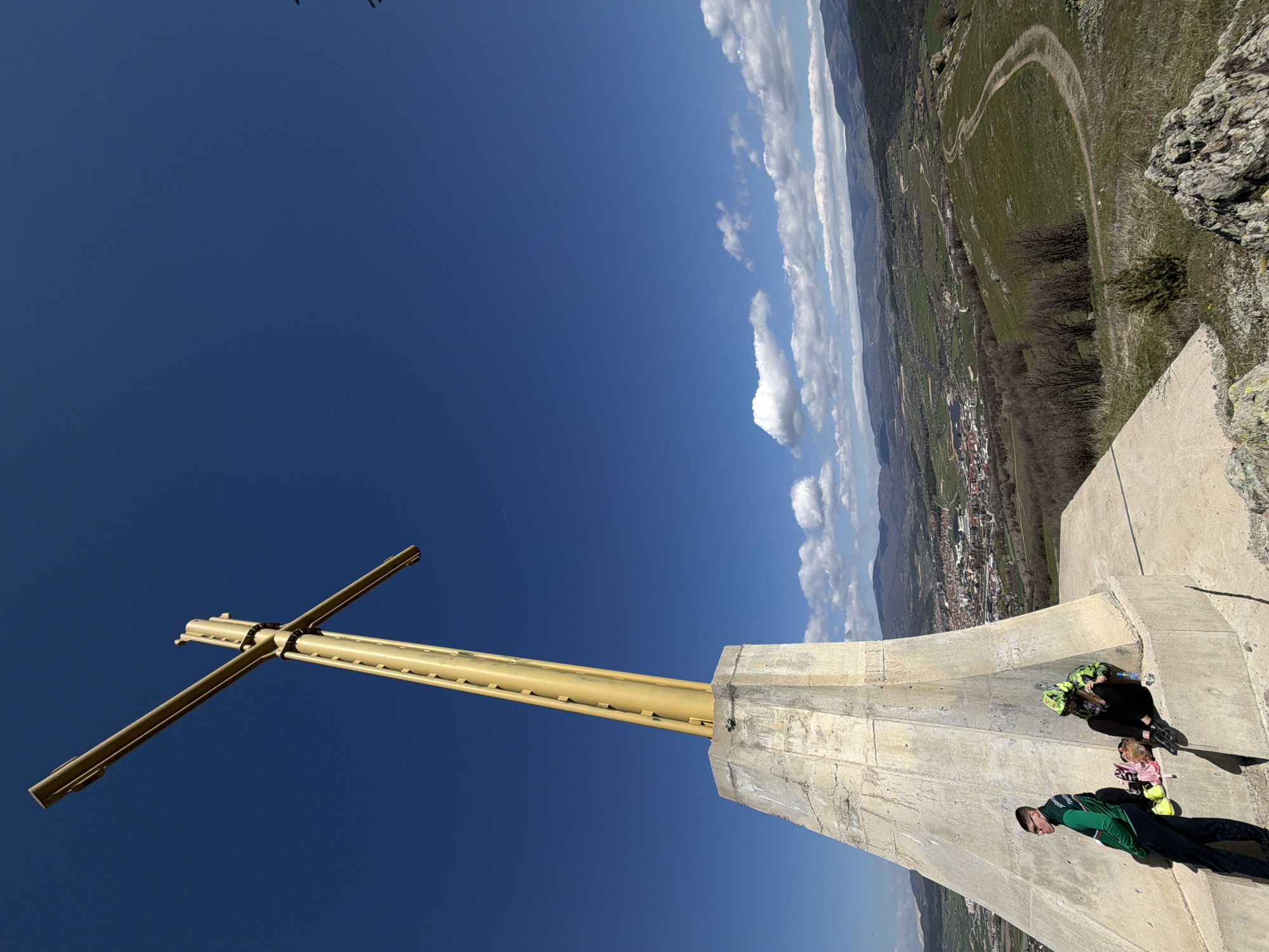 The image captures a high-angle view from the base of a large, yellow cross atop a concrete structure. Below, a town is visible in the distance. Two people are seated on the structure's edge. The sky is blue with scattered clouds. The location is Bata, Bulgaria.