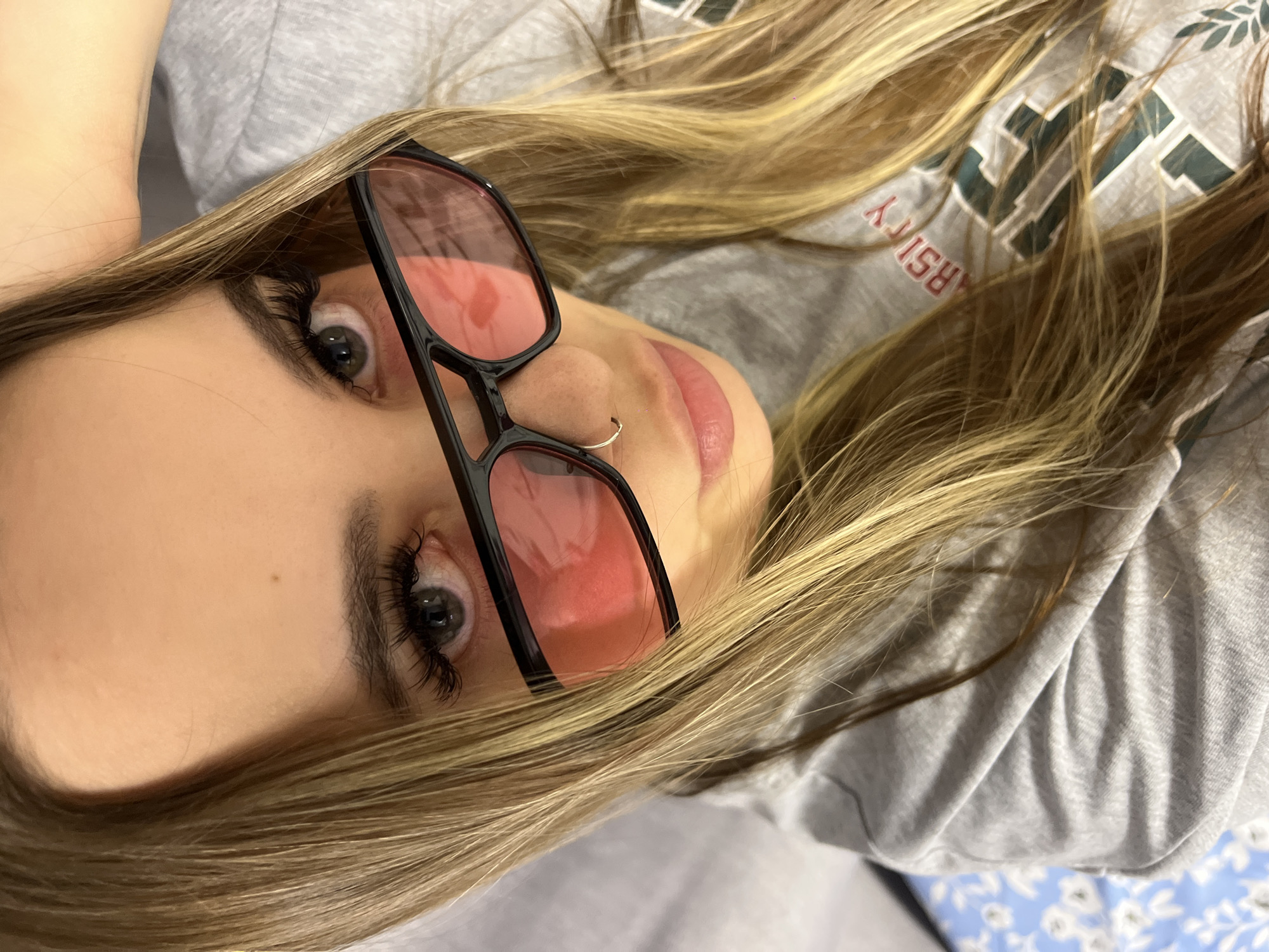 A woman with long blonde hair, wearing black-framed pink-tinted glasses and a light grey sweatshirt with "Varsity" printed on it, is lying down. She has a nose piercing. The background includes a blue and white floral-patterned surface. The location is not evident from the image.