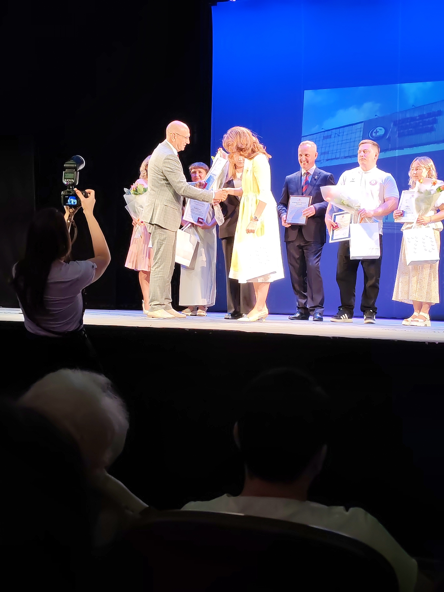 This image captures an award ceremony on a stage in the city of Chaikovsky, Russia. On the stage, a man in a gray suit is presenting a certificate to a woman in a yellow dress. Behind them, several other individuals stand, some holding bouquets of flowers and certificates. The backdrop of the stage features a large screen displaying an image of a modern building, possibly an industrial facility. In the foreground, the back of a person holding a camera is visible, suggesting a photographer documenting the event. The lighting indicates an indoor event, likely a theater or hall, with spotlights illuminating the stage and the audience in partial darkness.