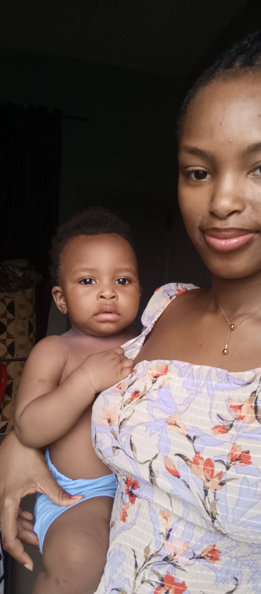 A close-up, brightly lit portrait features a woman holding a baby in an indoor setting in Keffi, Nigeria. The woman's face is partially visible on the right side of the frame, with her eyes looking towards the camera and a neutral expression. She has dark skin and her hair appears pulled back. She is wearing a light-colored, possibly white or light blue, floral patterned top with smocked detailing and thin straps. A delicate gold necklace with two spherical pendants rests on her chest. Her left arm is visible as she holds the baby.
The baby, an infant or young toddler, is positioned in the center-left, with their full face looking directly at the camera, displaying a neutral yet curious expression. The baby has dark skin and short, dark, curly hair. The baby's upper body is bare, revealing their chest and arms, and they are wearing light blue diaper cover or shorts. The baby's right hand is resting gently on the woman's shoulder strap.
The background is dark and out of focus, suggesting low light conditions or a dark wall behind the subjects. Faint patterns of a fabric or mat are visible on the left side, along with a dark curtain further back. The lighting appears natural, illuminating the subjects prominently against the darker backdrop, creating a sense of intimacy. No text is visible in the image.