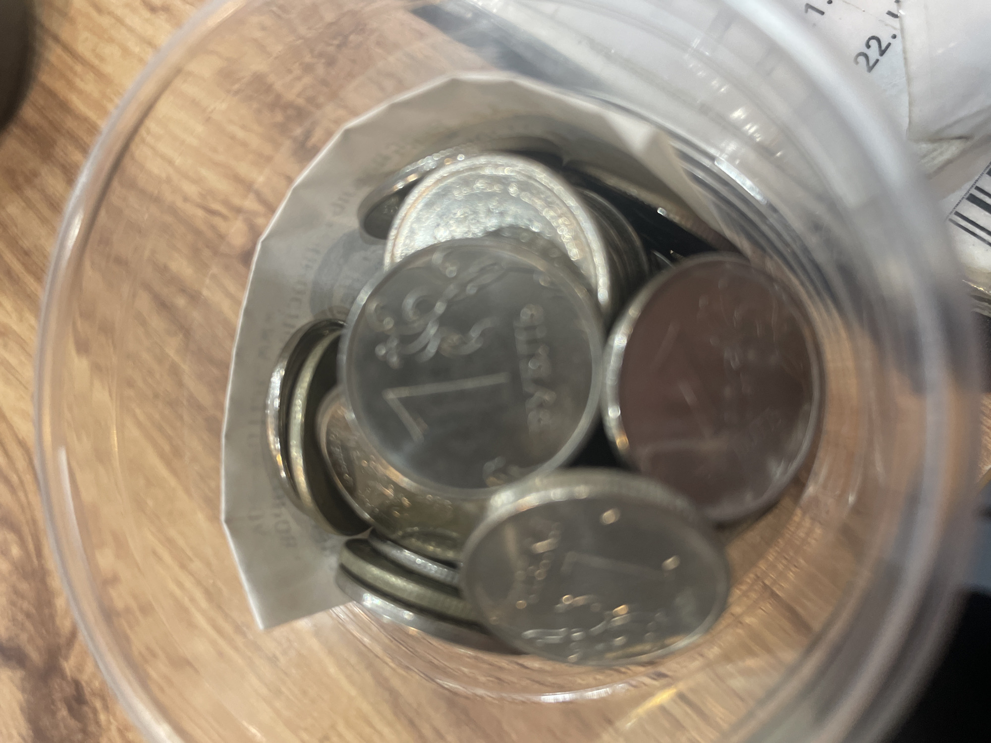 A clear plastic container filled with Russian Ruble coins is placed on a wooden surface. The coins are a mix of silver and gold colors, with some showing clear markings and others being obscured by the angle and lighting. A white paper receipt or label is partially visible inside the container, tucked between the coins. The background suggests an indoor setting with a wooden table or countertop. The image is slightly out of focus, but the general contents and arrangement are discernible. There are no people or animals present in the image. The scene is static and does not depict any action. The location is stated to be the city of Zhukovo, Russia.