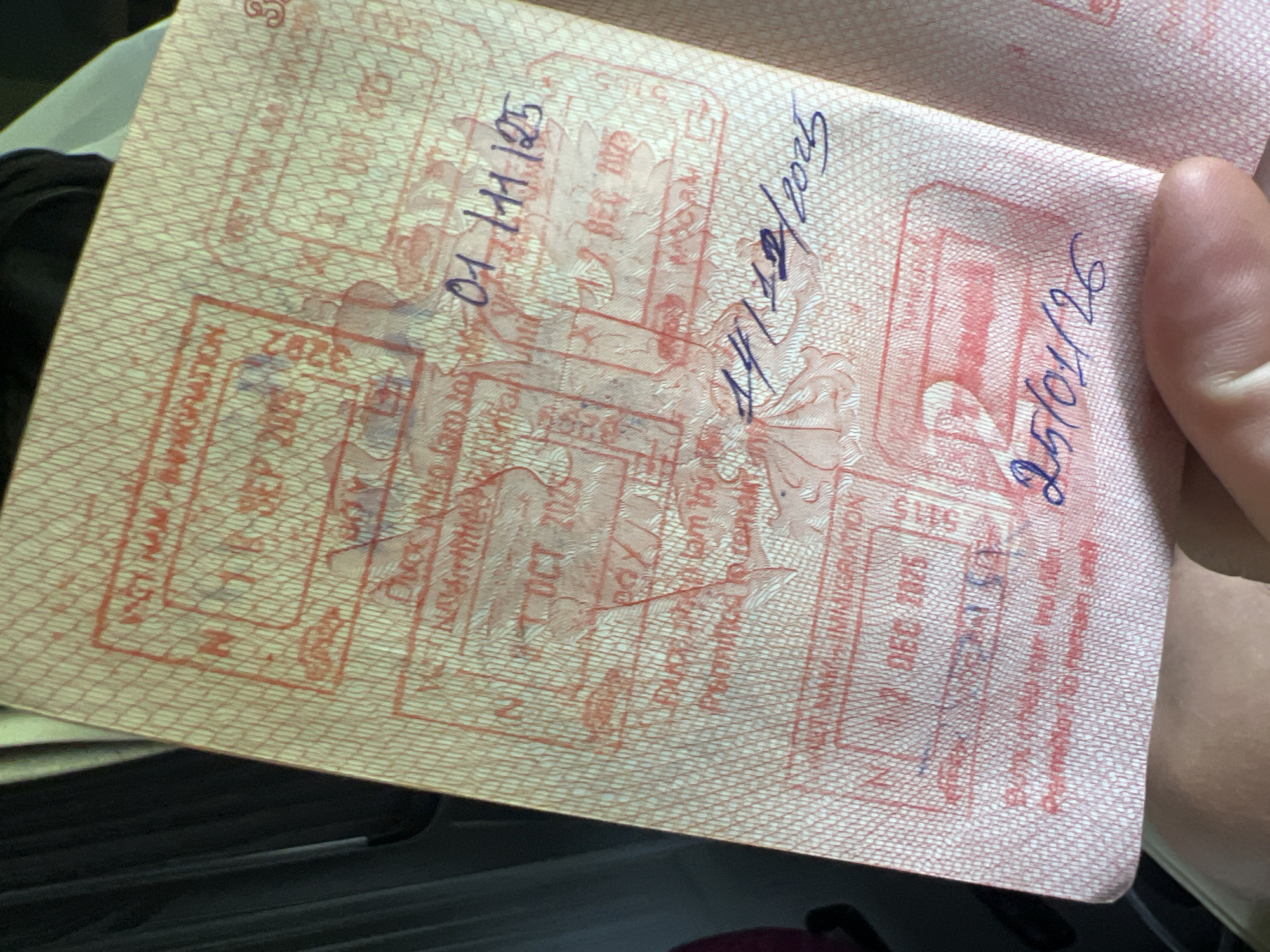 A passport page is angled with multiple entry/exit stamps and handwritten dates in blue ink. The stamps are in red ink and appear faded and partially obscured. The handwritten dates are "01/11/25", "11/12/2025" and "25/01/26". The stamp on the left contains "3282" and "SEP 2015", and below is "NA MIGRATION A". Another stamp contains "1 OCT 2015" and "Permitted to remain until". Another stamp contains "5115" and "11 DEC 2025" and "NA MIGRATION A". The location "Kon Ra Kiah, Vietnam" is not legible in this image. A human hand is partially visible holding the passport. The environment appears to be dimly lit.