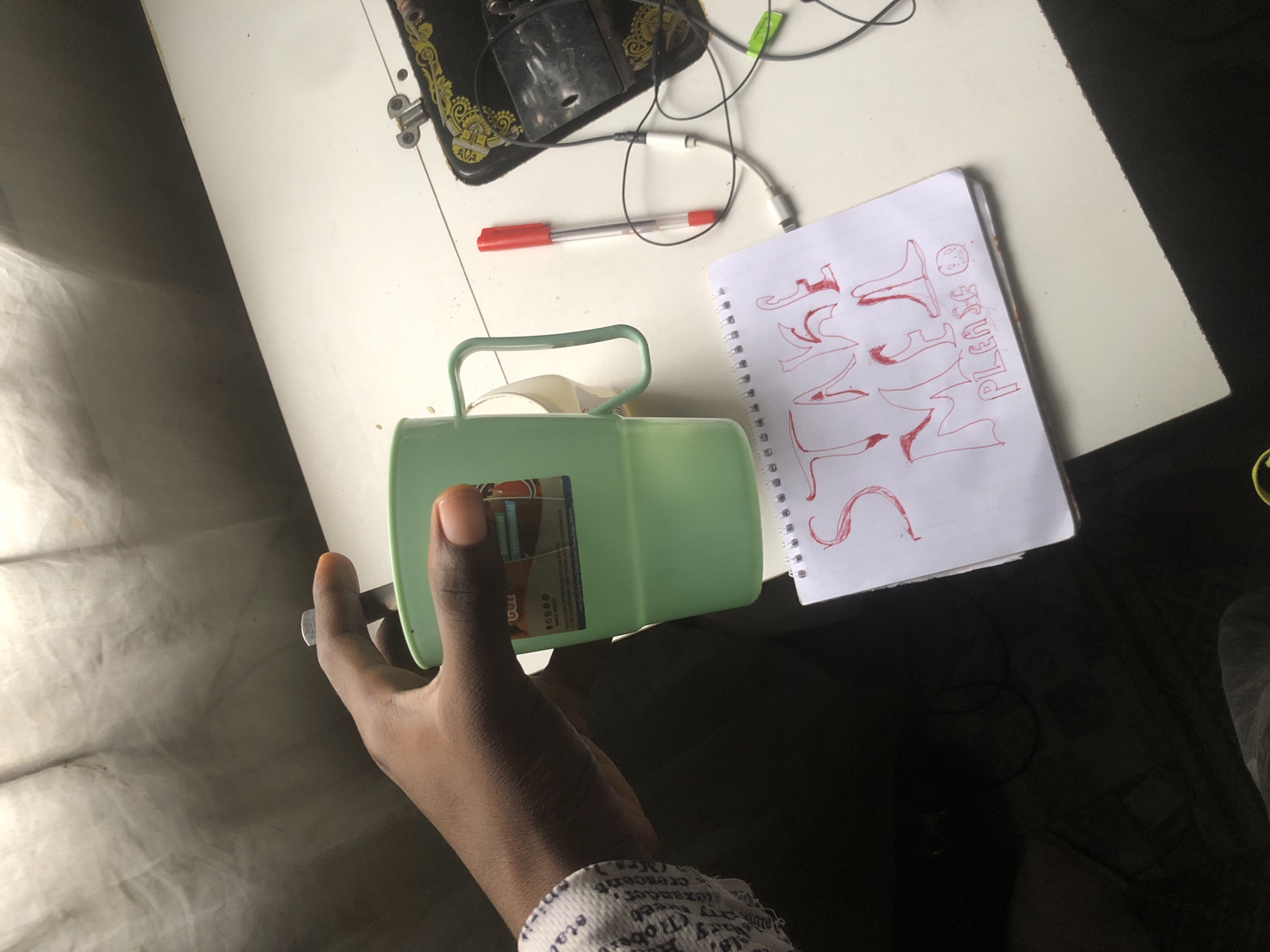 A close-up, overhead shot shows a person's right hand, with dark skin and manicured orange nails, holding a light green plastic mug with a white interior and a handle. The mug contains a white object, possibly a lid or another cup. The hand is positioned on a white surface that appears to be a table. To the right of the mug is a white notepad with a spiral binding, displaying a message written in red marker that reads "TAKE ME PLEASE" with a smiley face. Next to the notepad is a red and clear plastic pen. A sewing machine, partially visible on the right side of the frame, has black and gold ornate detailing and is connected to wires. The background is a draped curtain or fabric, with light filtering through, creating shadows and a soft, diffused illumination. The overall scene suggests a domestic or workshop environment, possibly in Ado, Nigeria, given the context provided.