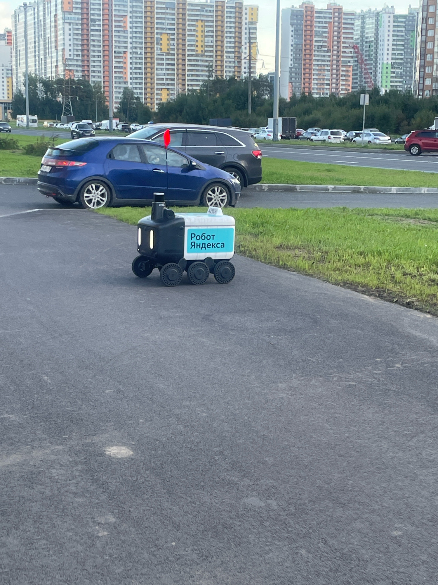 The image shows a delivery robot with the inscription "Робот Яндекса" (Yandex Robot) on its side, parked on an asphalt surface. In the background, there are several parked cars, including a blue hatchback and a dark SUV. Behind the cars, a grassy area leads to a road with more vehicles. Further in the background, numerous tall residential buildings are visible, characteristic of a large urban area. A red flag is attached to the antenna of the blue car. The overall scene appears to be a street or parking area in a modern residential district of Saint Petersburg, Russia, possibly during daylight hours under a slightly overcast sky.