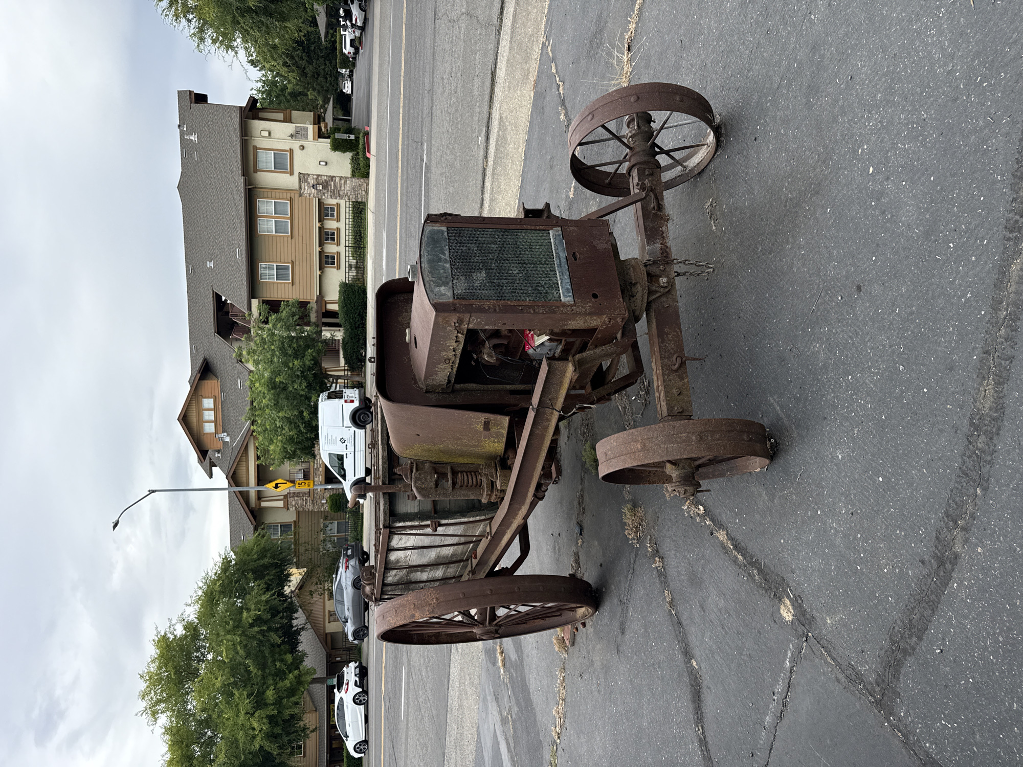 The image depicts an abandoned, rusted tractor with wooden sideboards, positioned on an asphalt surface in Fair Oaks, California. The tractor is centrally located in the frame and appears to be a historical artifact. In the background, a modern, multi-story apartment complex is visible, along with trees and a street with several vehicles, including a white van, a grey sedan, and two white cars. The sky is overcast, contributing to a muted lighting condition. The scene is set outdoors, likely in a residential or commercial area of Fair Oaks, United States.